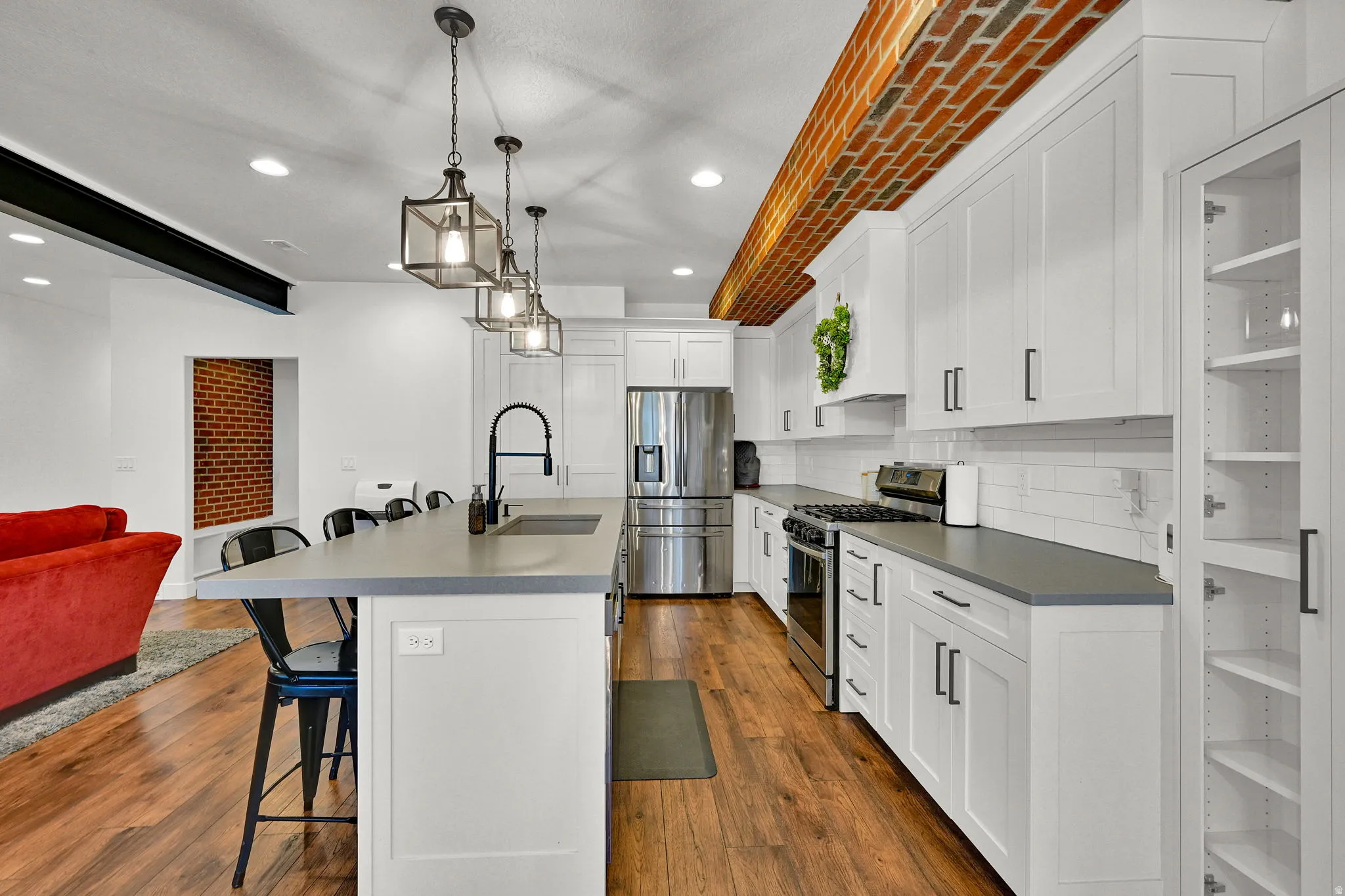 Open-concept kitchen with wood-finish flooring, white shaker cabinetry, stainless steel appliances, a center island with sink, and a brick-accented ceiling feature