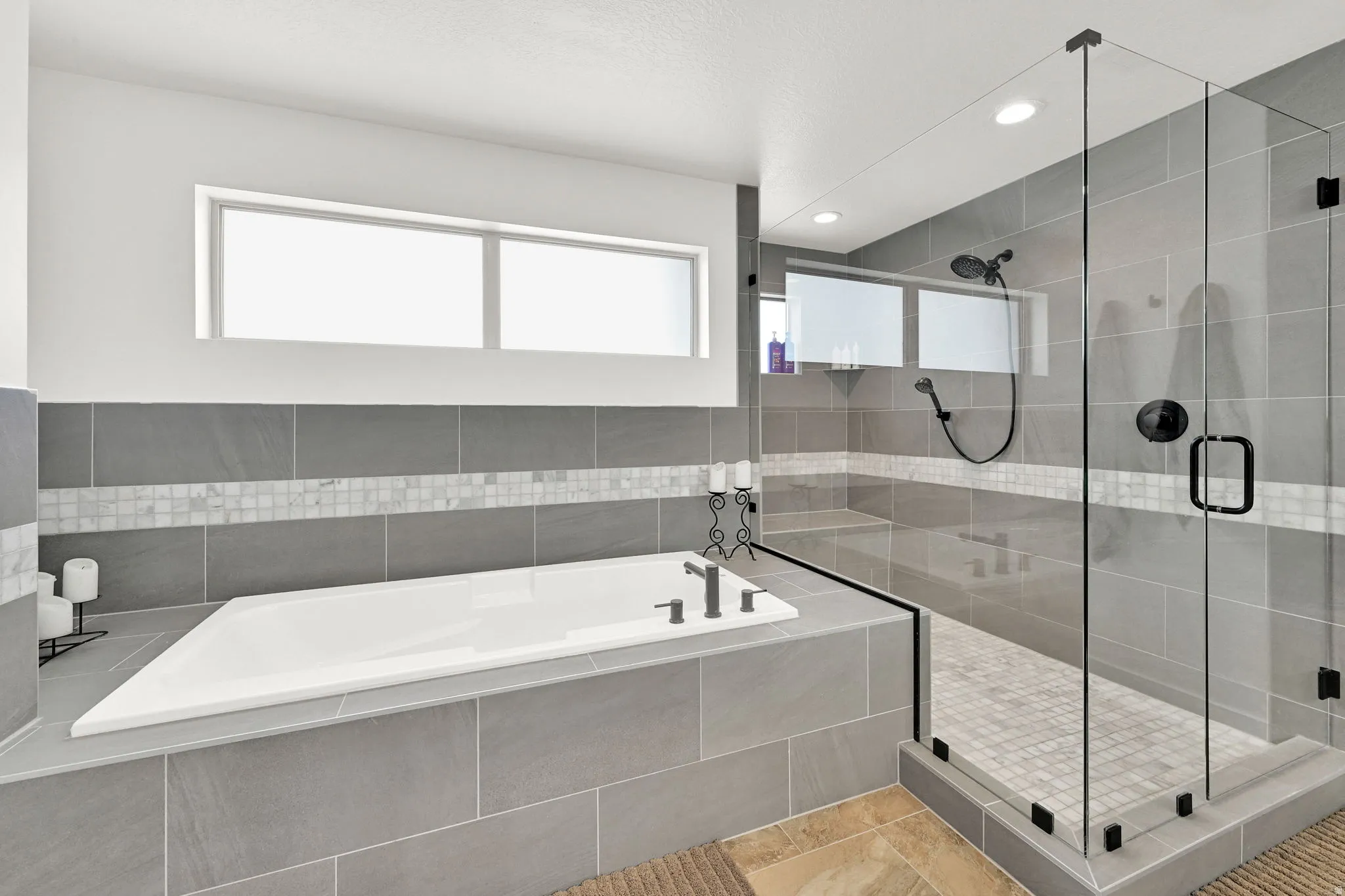 Soaking tub with dark fixtures set in grey tile surround, complemented by a glass-enclosed shower with matching grey tile walls and mosaic accent