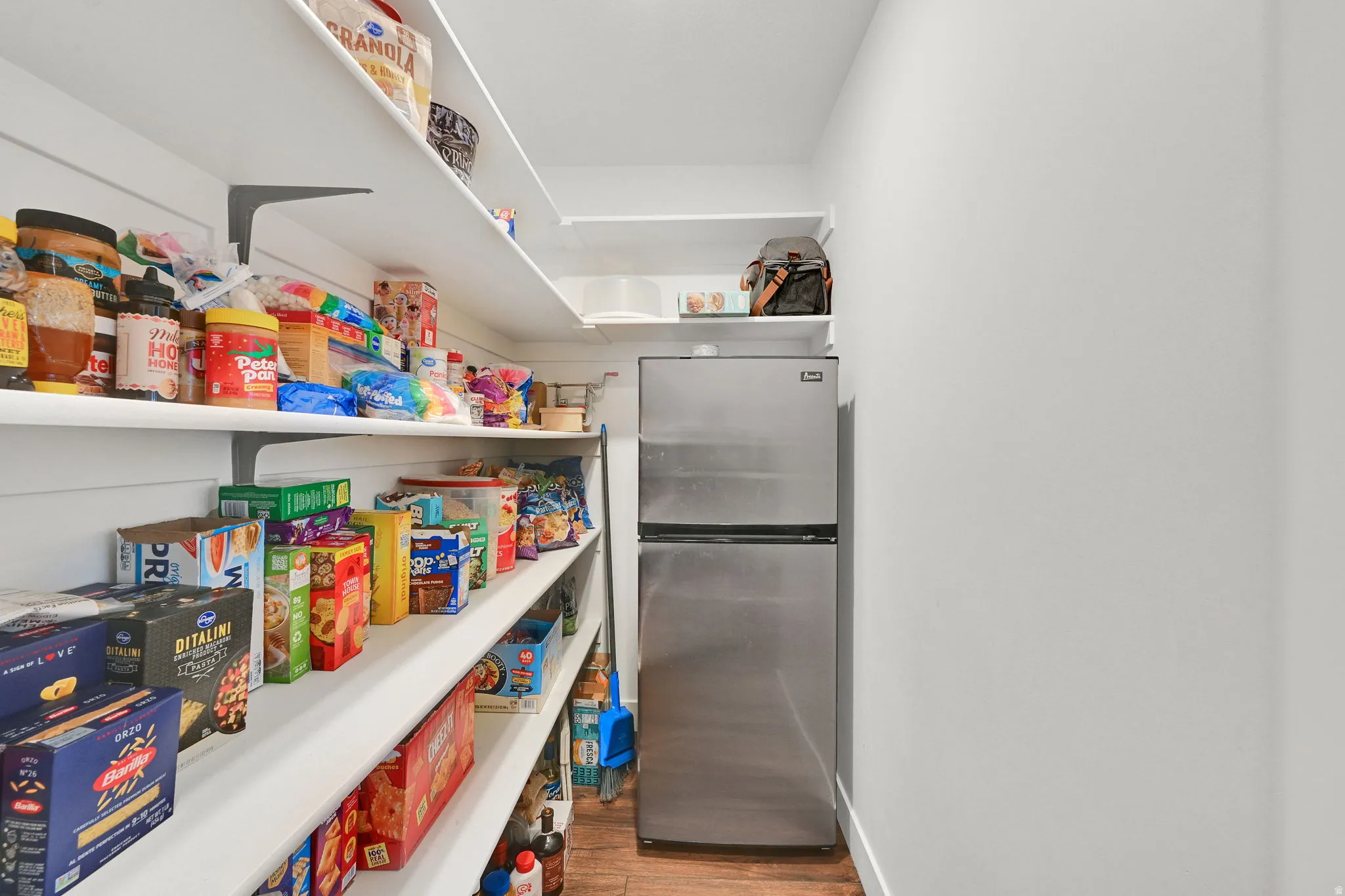 Walk-in pantry featuring extensive white shelving, wood-finish flooring, and a stainless steel refrigerator