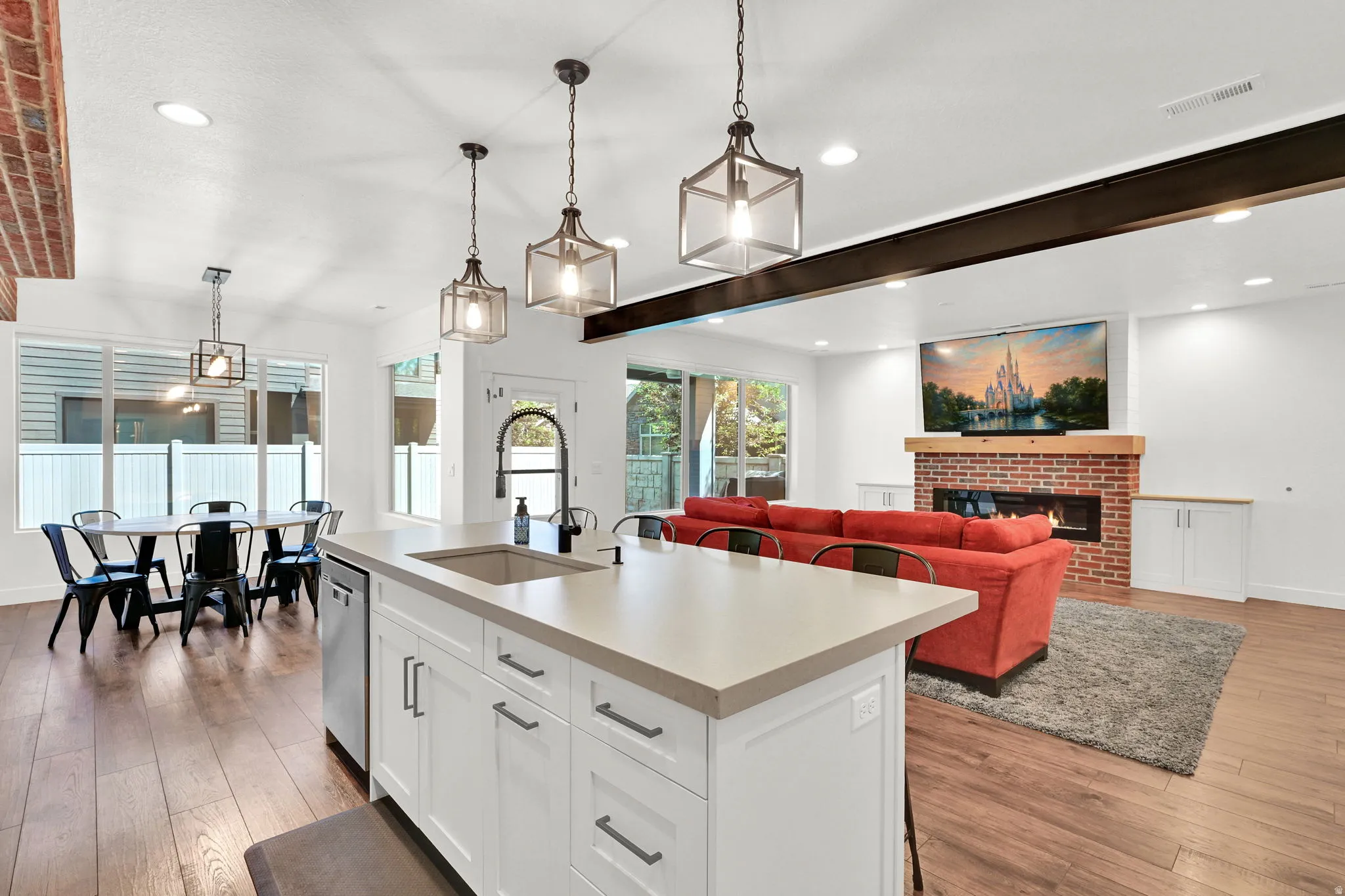Open-concept living space featuring wood-finish flooring, a kitchen island with integrated sink, a brick fireplace with wood mantel, and multiple large windows