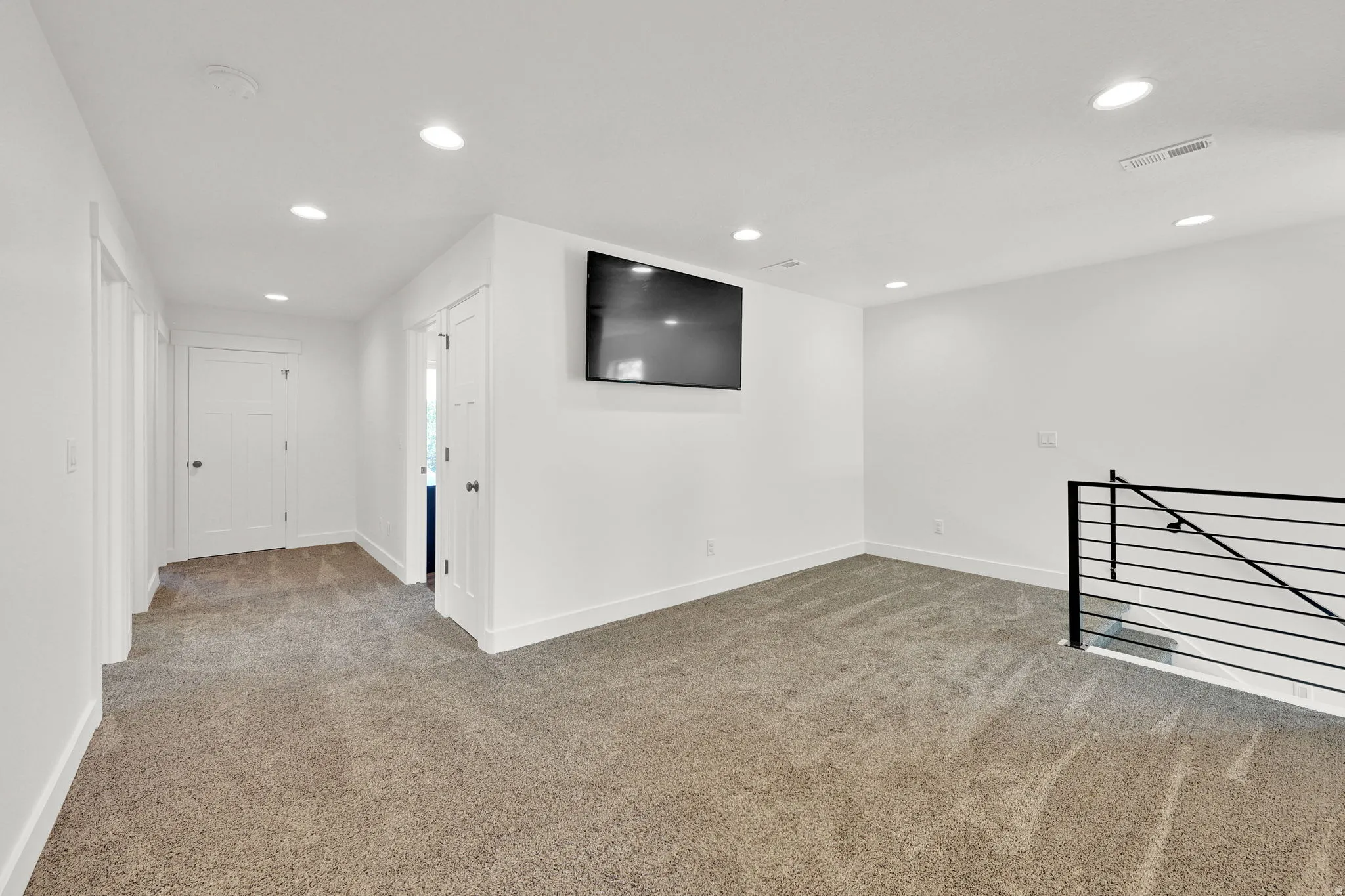 Carpeted loft area featuring recessed lighting, a mounted display, white walls, and a contemporary black metal railing