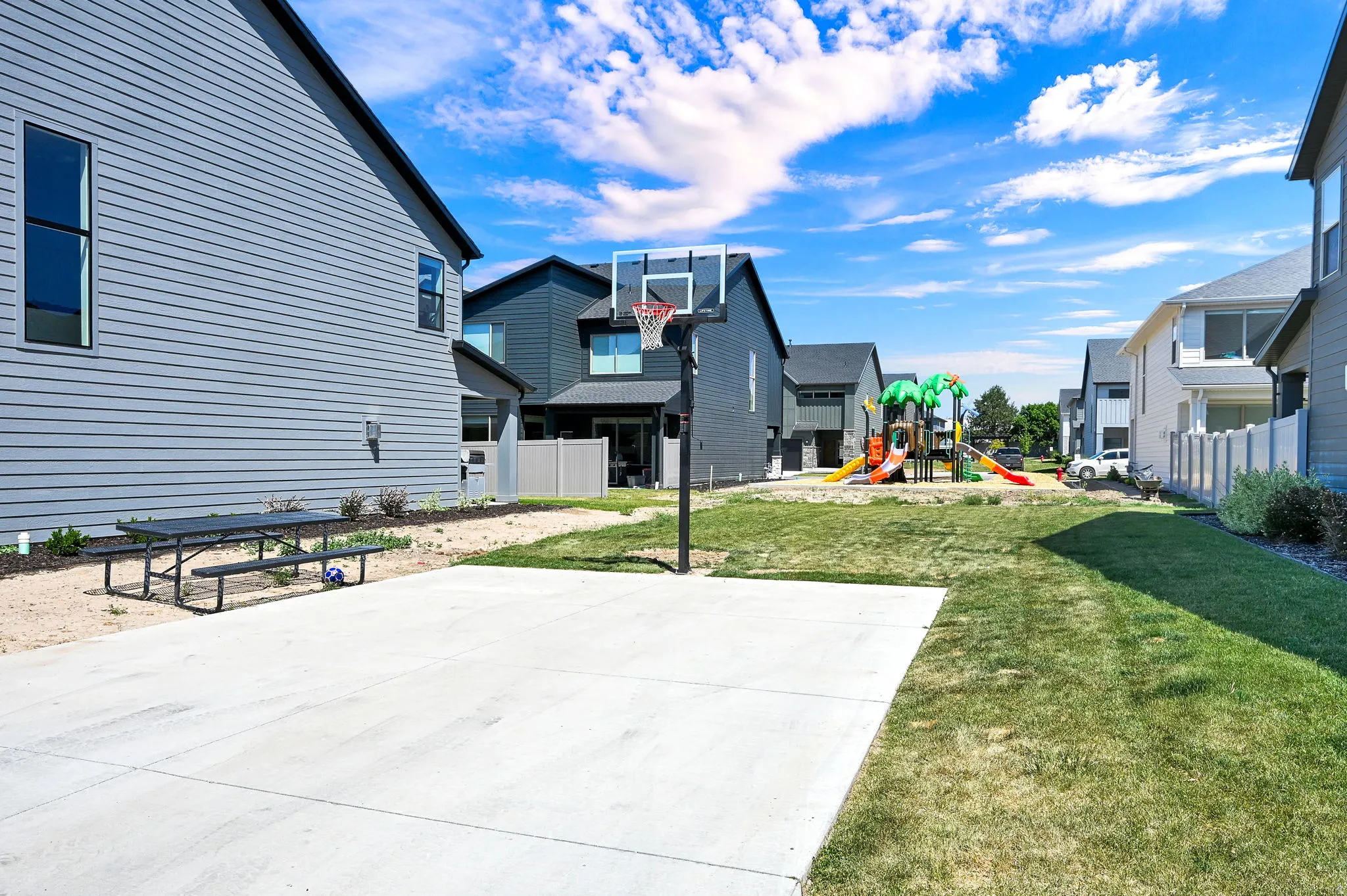 Outdoor amenity space featuring a concrete pad with a basketball hoop, an adjacent grassy area, and a playground structure in the distance