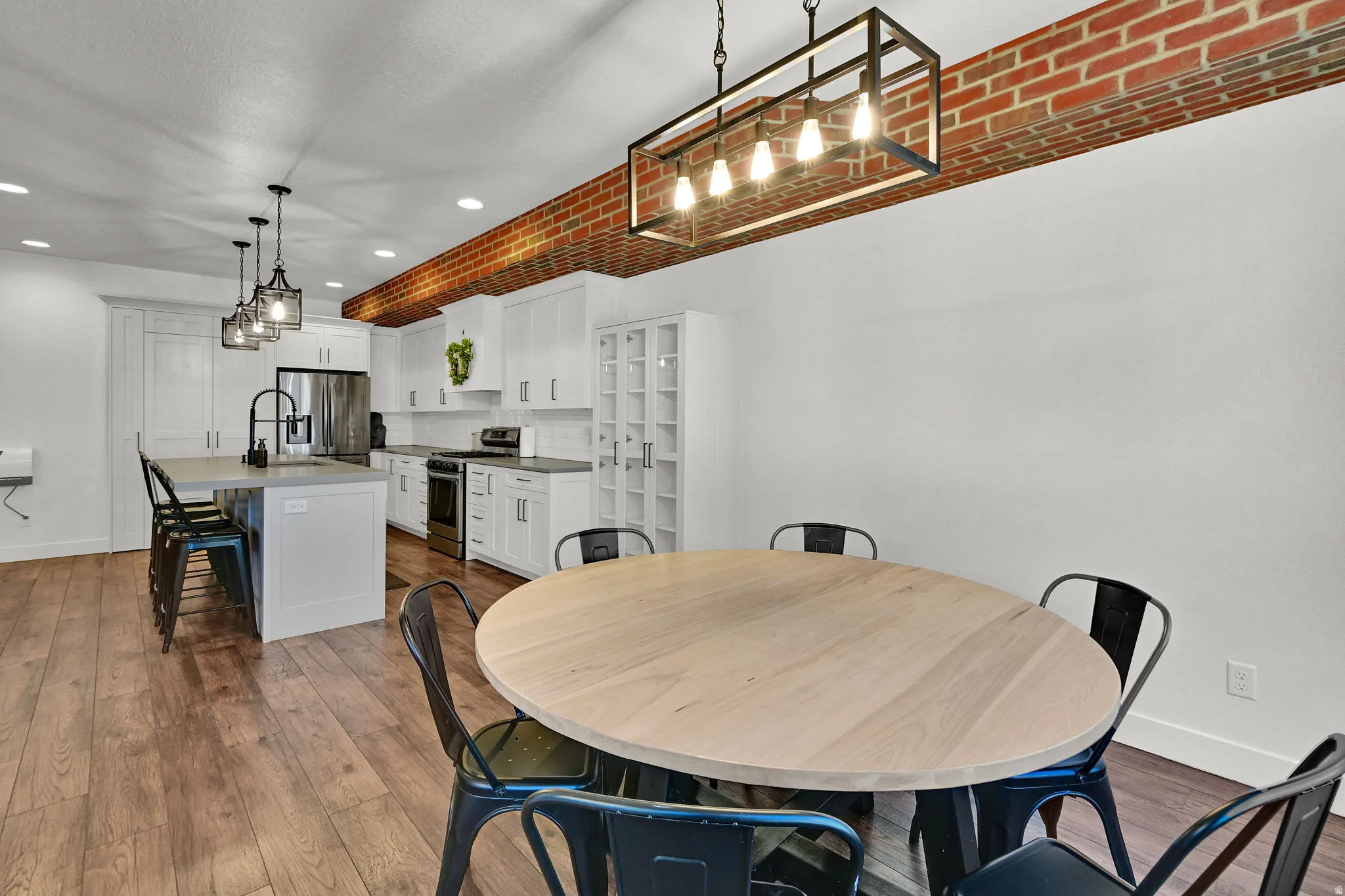 Open-concept kitchen and dining area featuring wood-finish flooring, white cabinetry, a kitchen island with seating, a stainless steel refrigerator, and a brick-accented ceiling