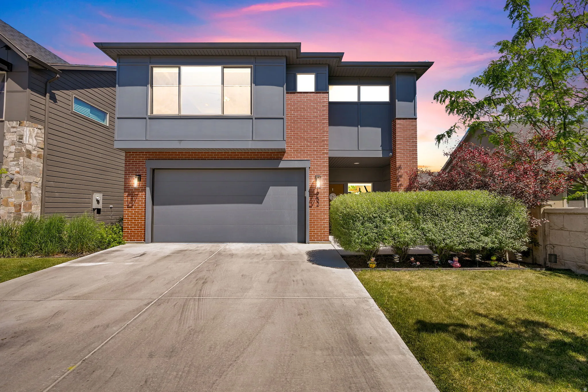 Contemporary exterior featuring brick and gray paneling, an attached garage, a concrete driveway, a manicured lawn, and mature landscaping