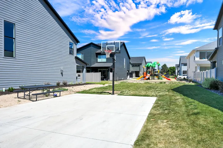 Outdoor amenity space featuring a concrete pad with a basketball hoop, an adjacent grassy area, and a playground structure in the distance