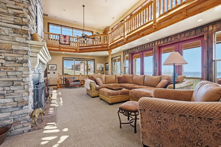 Spacious living area featuring a floor-to-ceiling stone fireplace, high ceilings, an upper-level balcony with wood railing, expansive windows, and light-colored carpeting