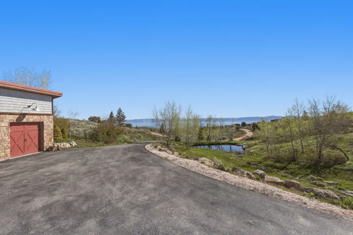 Asphalt driveway curving through a natural landscape featuring a stone and shingle-sided structure with barn-style doors, a tranquil pond, and distant mountain-like formations