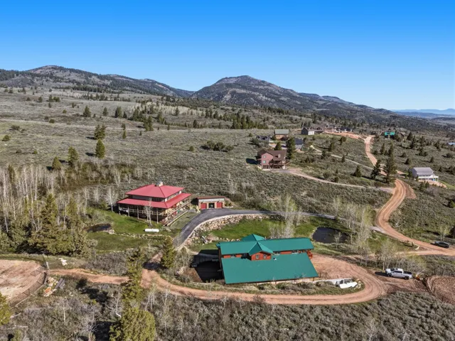 Rural property featuring a green-roofed residence, red-roofed residence with wrap-around porch, two-tone metal-roofed outbuilding, paved driveway, and natural landscaping