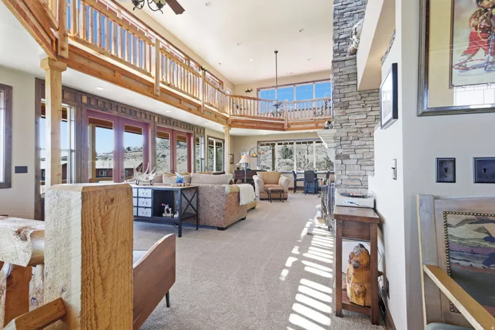 Great room featuring high ceilings, a stacked stone fireplace, a second-story interior balcony, exposed wood beams, and a wall of windows