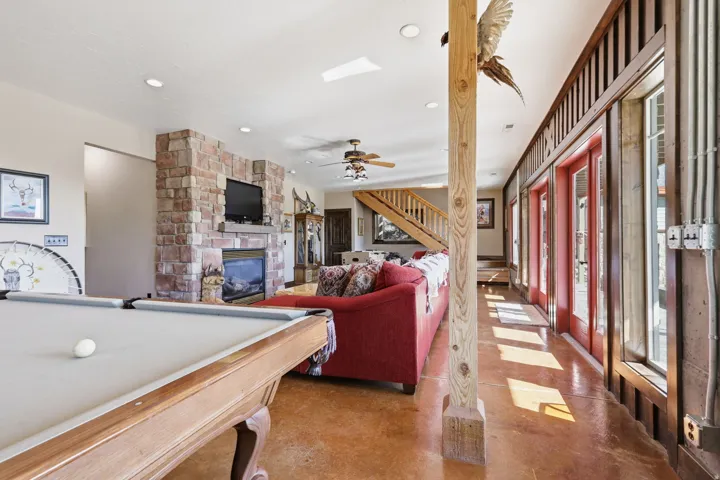 Spacious recreation area featuring a stacked stone fireplace, wood-finish flooring, recessed lighting, exposed wood ceiling beam, and a staircase with wood railings