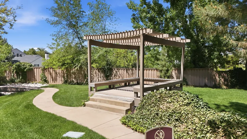 Expansive grassy area featuring a wood pergola with built-in seating, a winding concrete pathway, mature trees, and a wooden privacy fence
