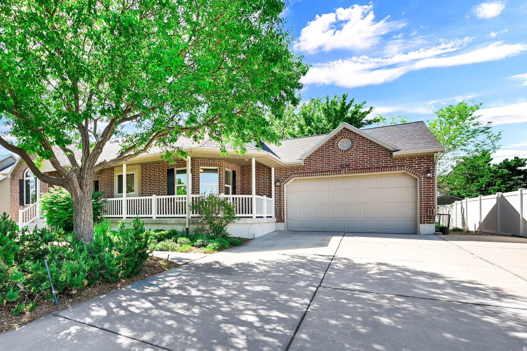 Brick exterior with a covered porch featuring white railings, an attached garage, a mature shade tree, and established landscaping
