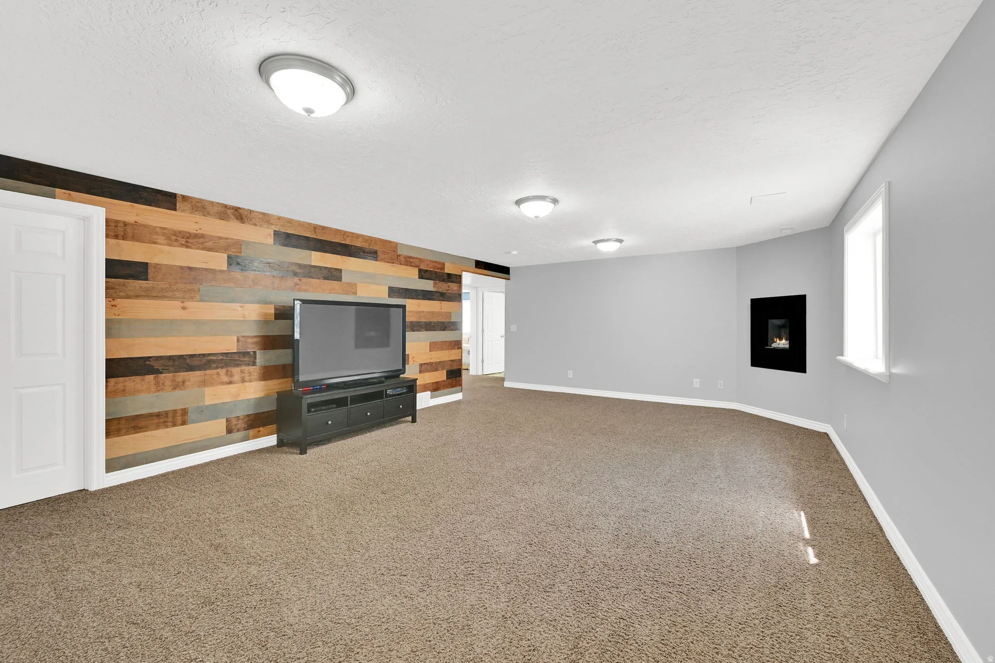 Spacious room featuring a multi-toned wood plank accent wall, wall-mounted fireplace, light gray painted walls, a window with white trim, and a textured carpeted floor