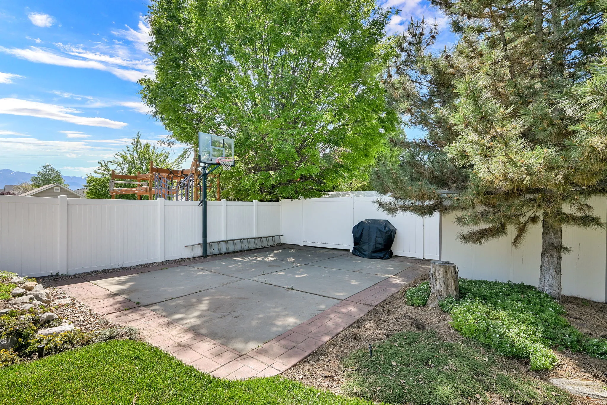 Backyard patio featuring a basketball hoop on a concrete slab, bordered by brick pavers