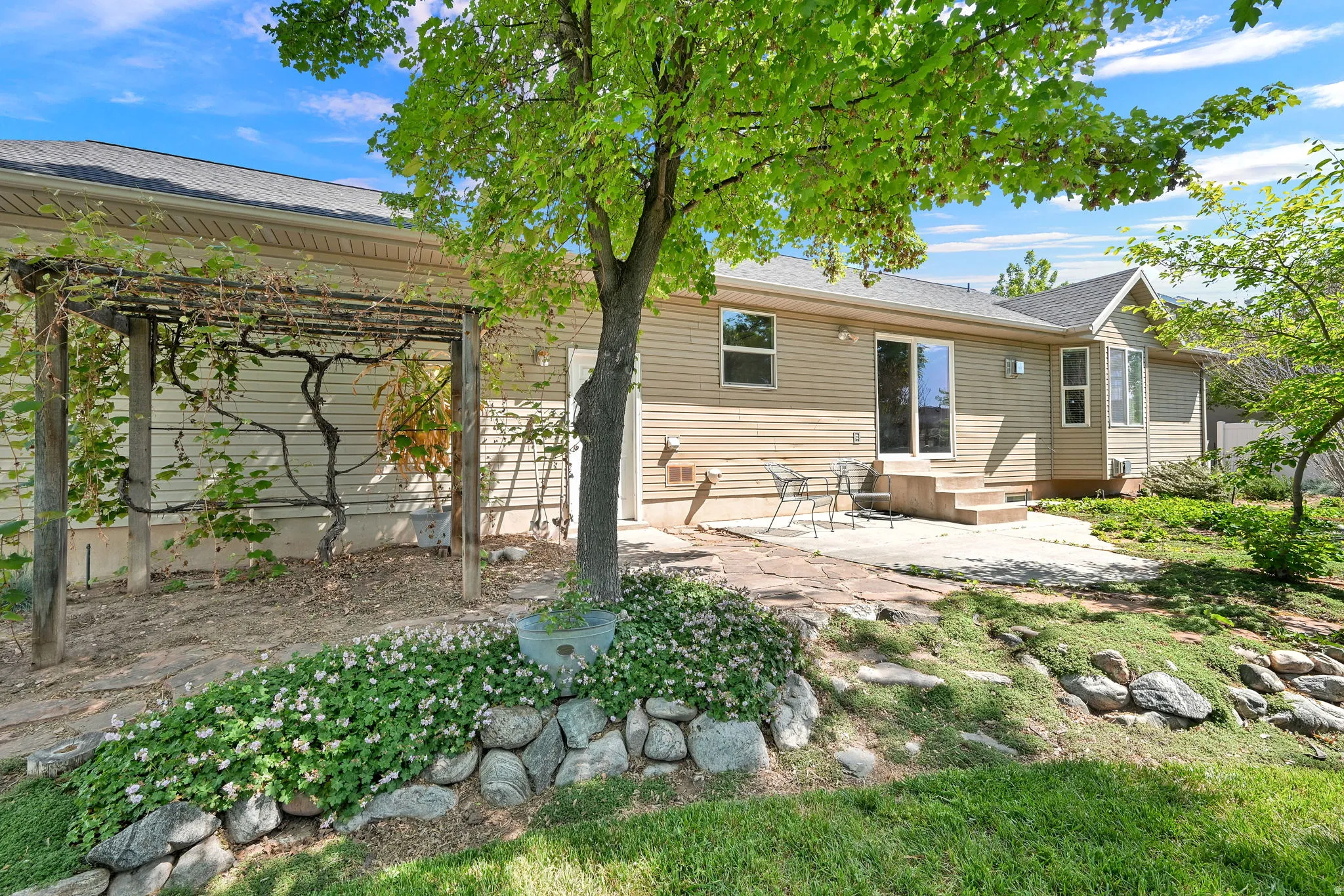 Rear exterior featuring a stone patio, sliding glass door entry, pergola with climbing vines, mature trees, and a stone retaining wall
