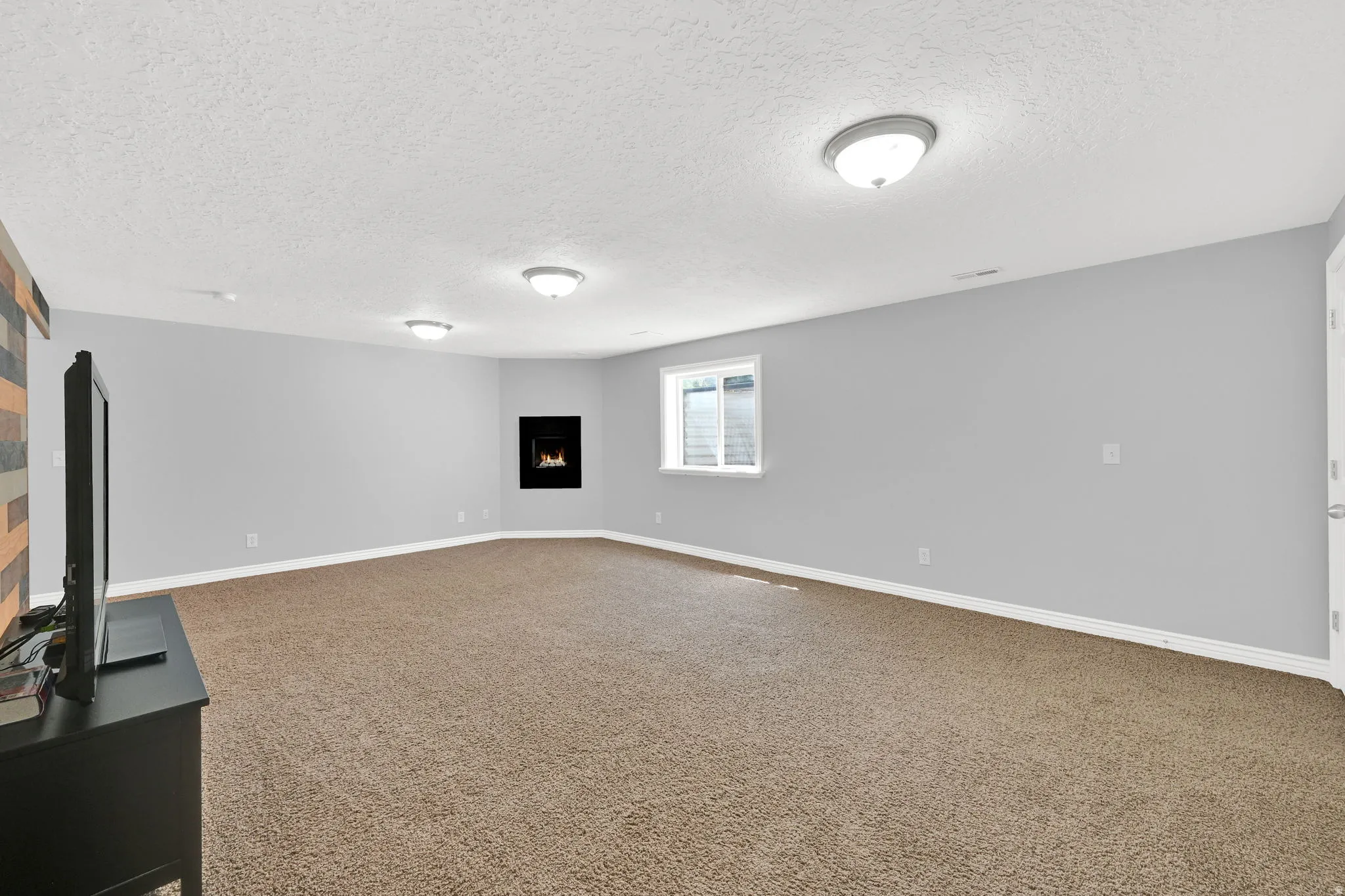 Spacious room featuring neutral carpeting, light gray walls, a built-in electric fireplace, and a window providing natural light