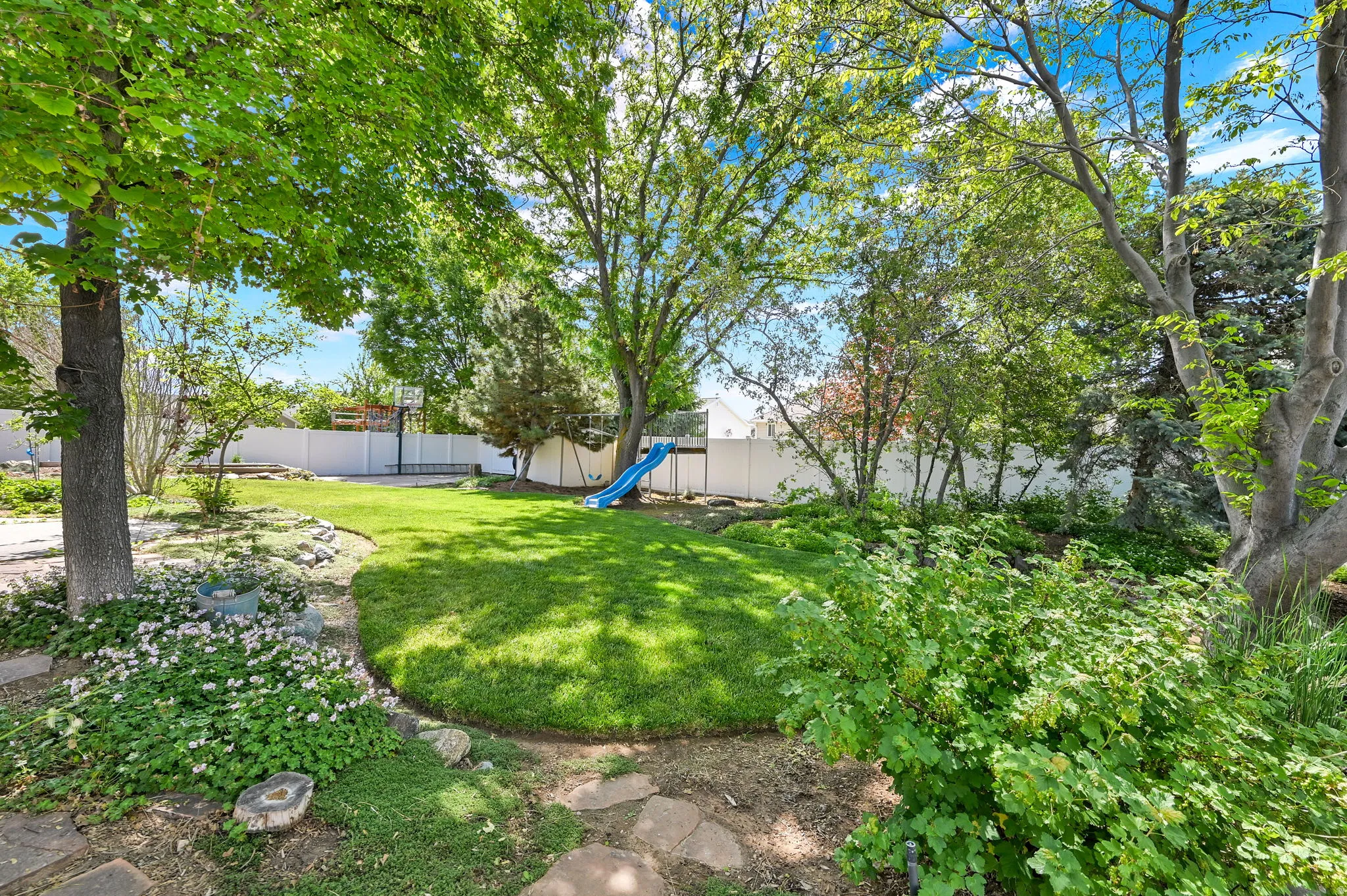 Spacious backyard featuring a manicured lawn, established landscaping, mature trees, and a white perimeter fence