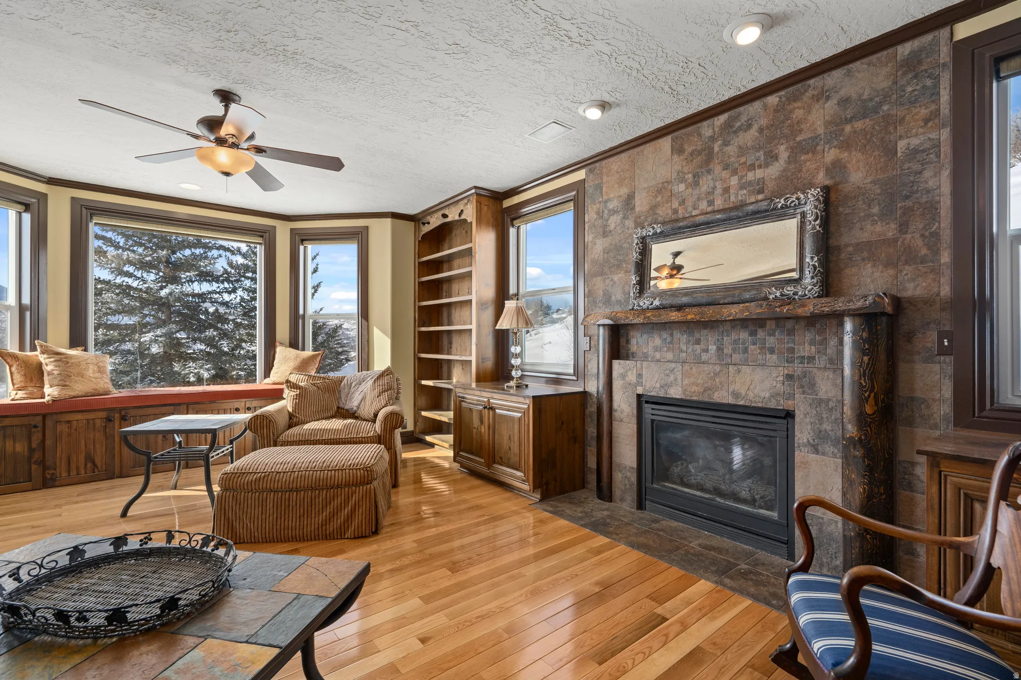 Hardwood flooring throughout a living space featuring a bay window with built-in storage, a ceiling fan, and a fireplace with a stone surround and mantel