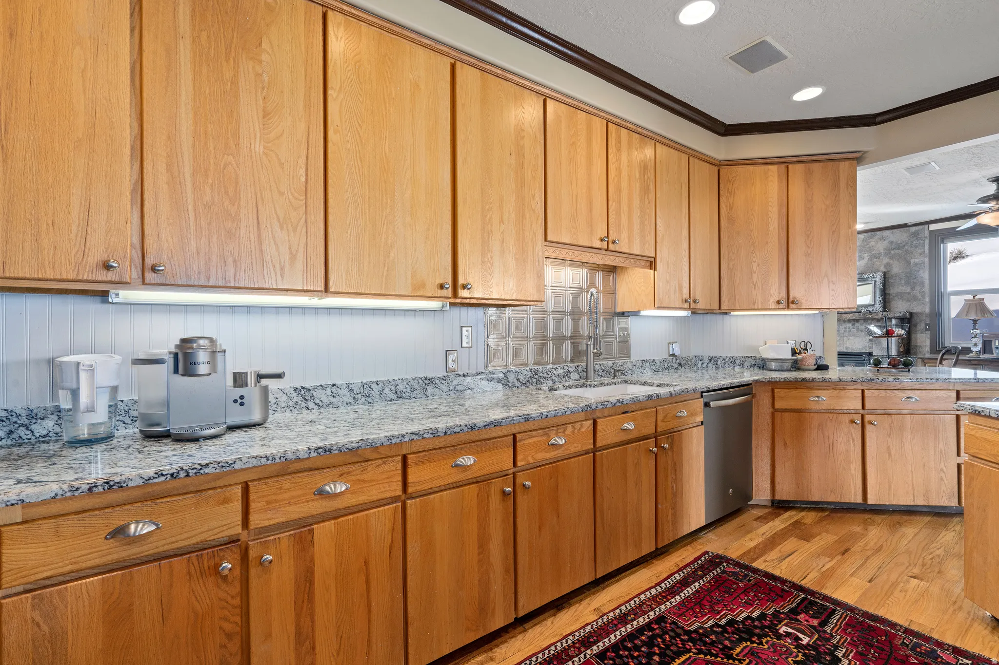 Kitchen featuring wood-finish cabinetry, light-toned countertops, tile backsplash, stainless steel dishwasher, and wood-finish flooring