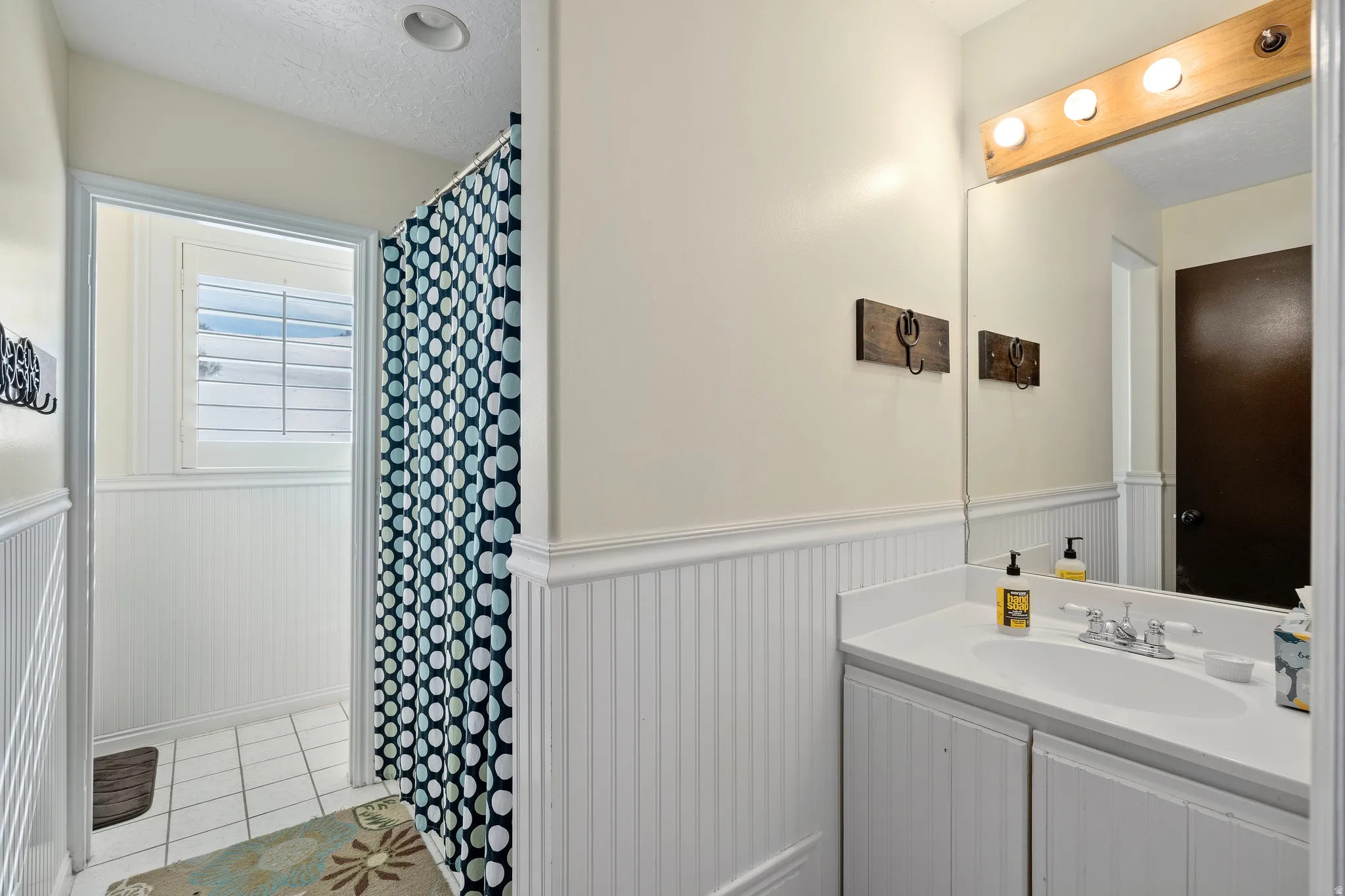 Bathroom featuring white beadboard paneling, a white vanity with integrated sink, a large wall-mounted mirror, and a shower area with white tile flooring and a shuttered window