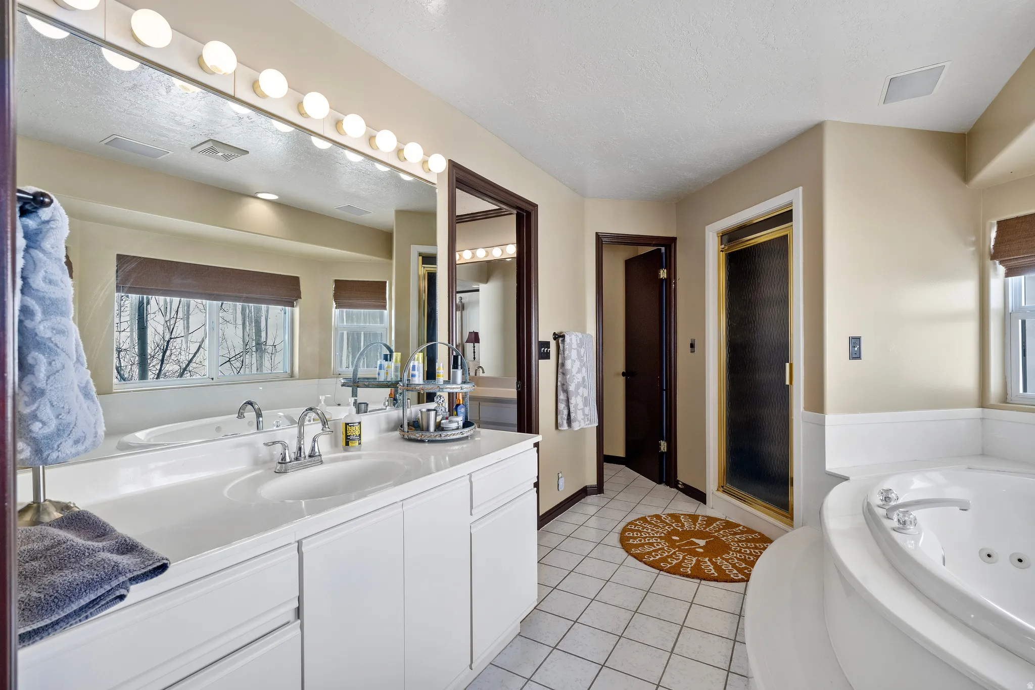 Bathroom featuring a double vanity with integrated sinks, a large mirror with sconce lighting, an enclosed shower with a gold-toned frame, a jetted soaking tub, and white tile flooring