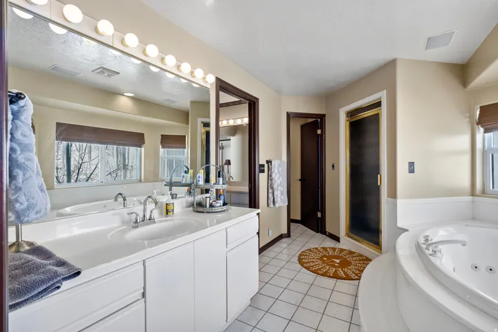 Bathroom featuring a double vanity with integrated sinks, a large mirror with sconce lighting, an enclosed shower with a gold-toned frame, a jetted soaking tub, and white tile flooring