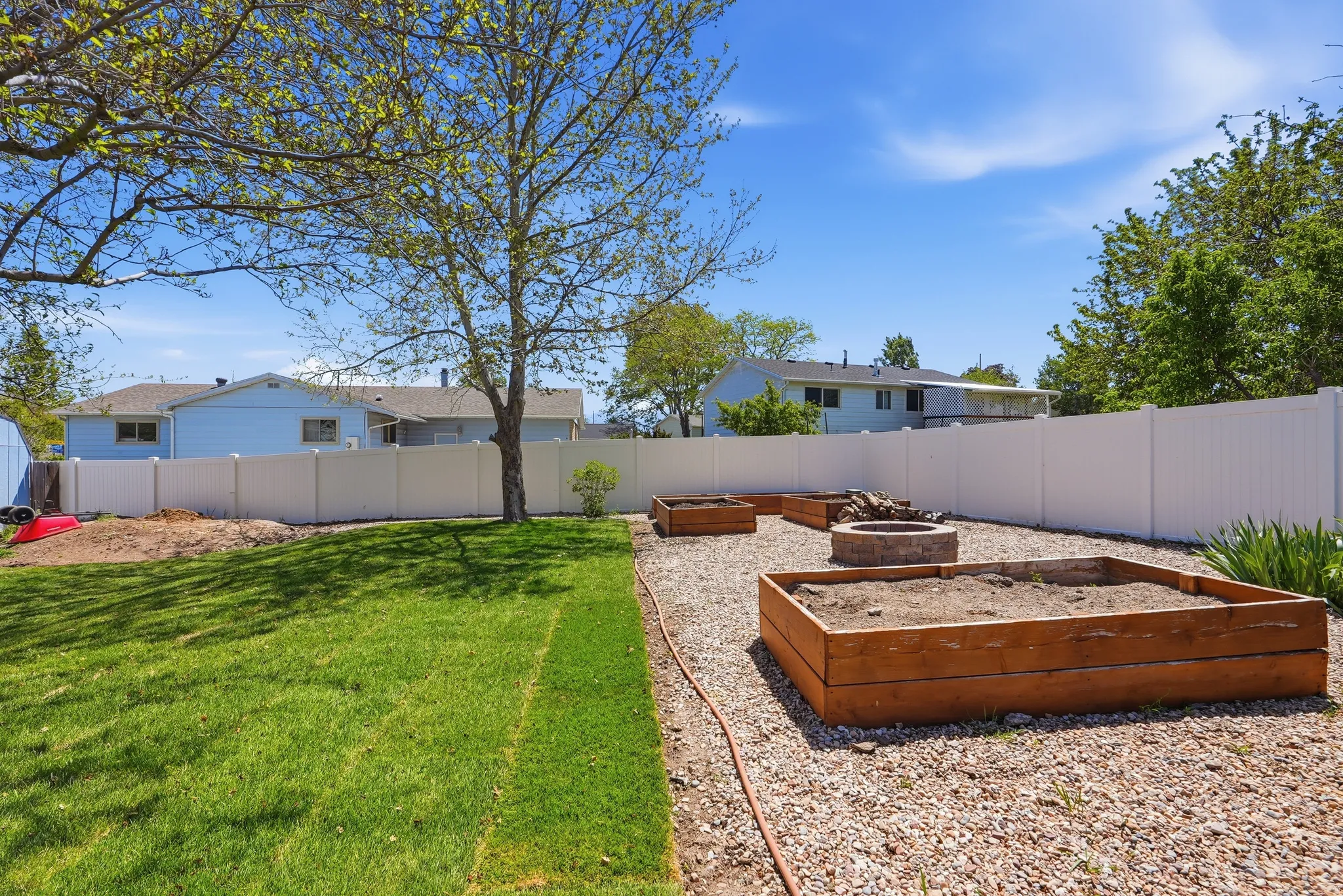 Spacious backyard featuring a manicured lawn, white privacy fencing, and a gravel area with multiple raised garden beds