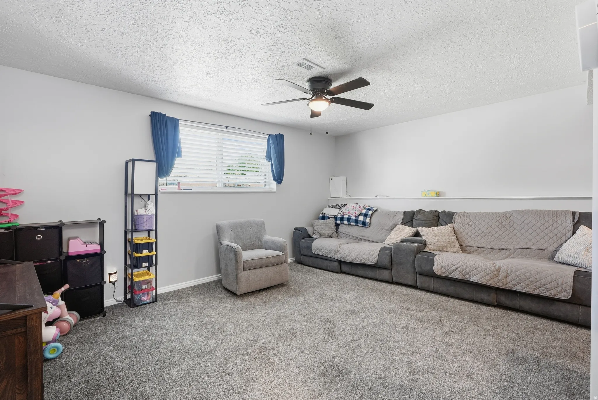 Spacious room featuring light gray walls, textured ceiling, a window with blinds, and a built-in ceiling fan with lighting