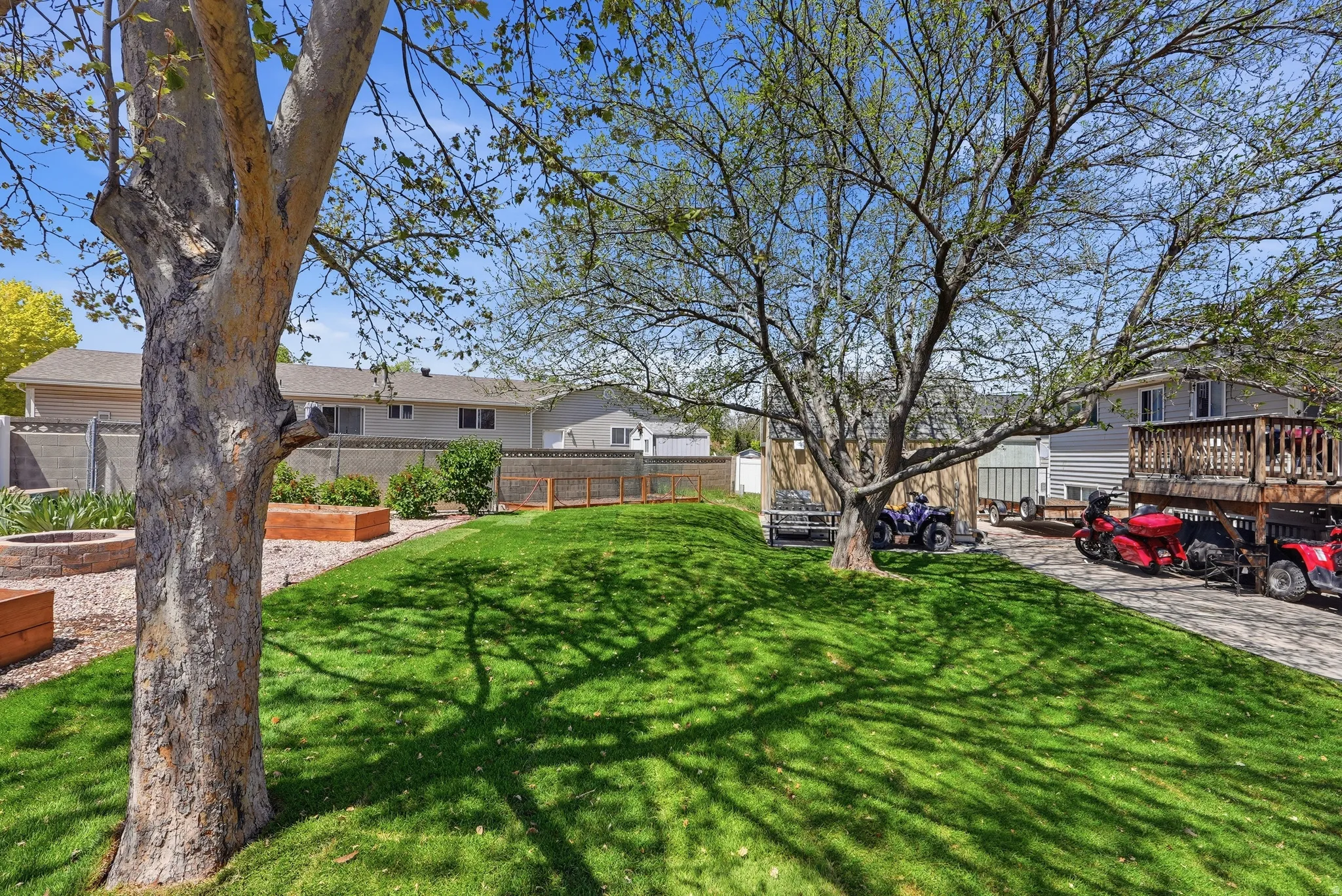 Expansive backyard featuring a lush lawn, mature trees, a built-in fire pit with stone surround, and terraced garden beds