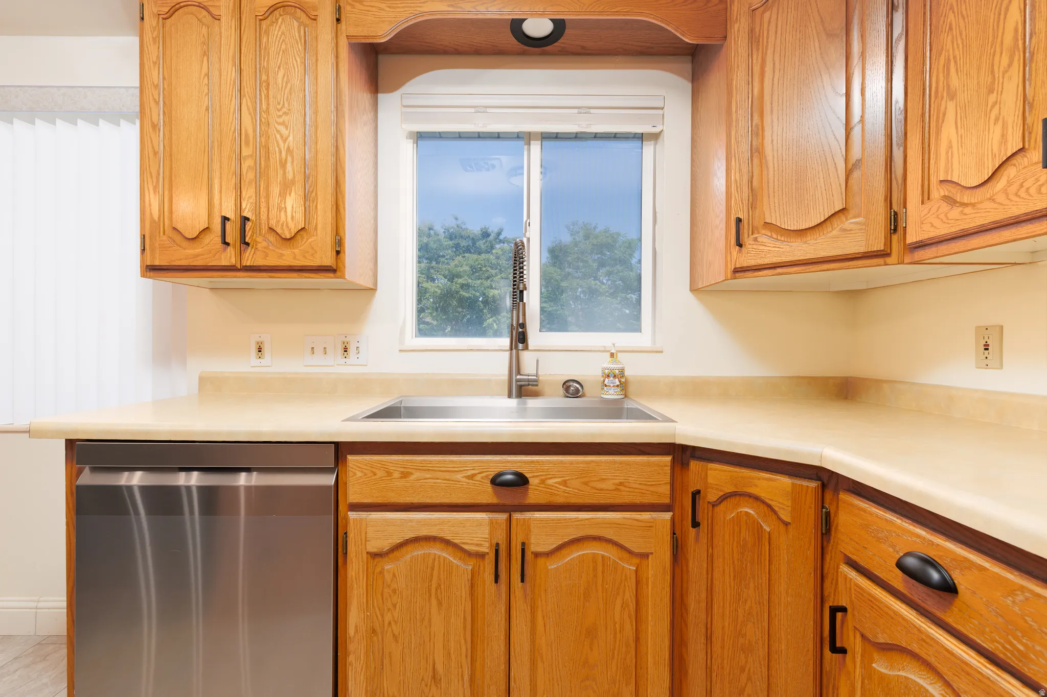 Kitchen featuring wood cabinetry, stainless steel dishwasher, undermount sink with a high-arc faucet, and light-toned countertops