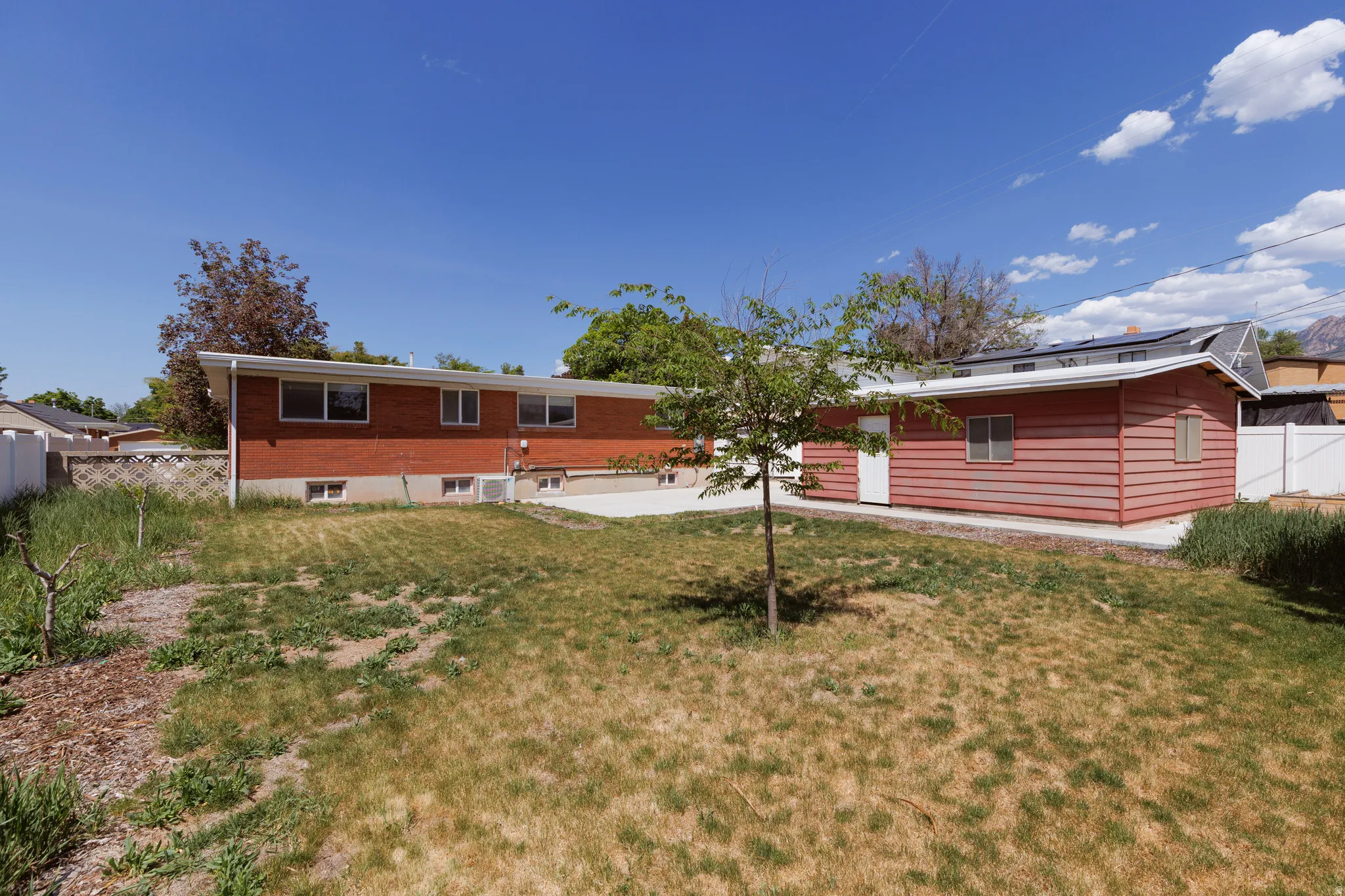 Expansive backyard featuring a red brick dwelling with multiple windows, a concrete patio area, and a separate structure with red horizontal siding