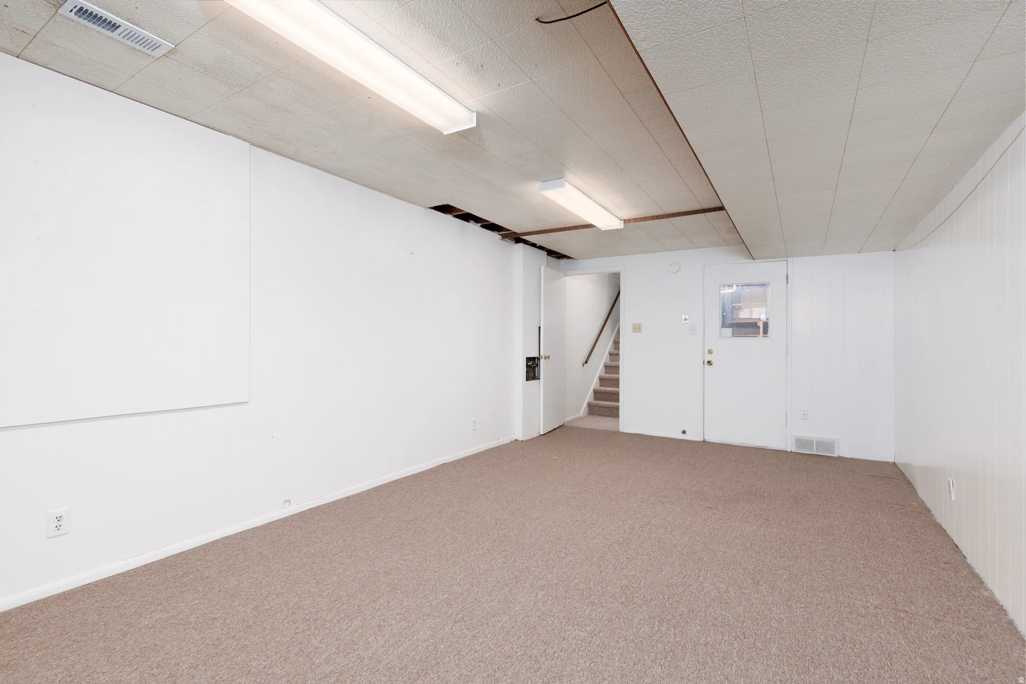 Spacious room featuring neutral carpeting, white walls, and a drop ceiling with fluorescent lighting