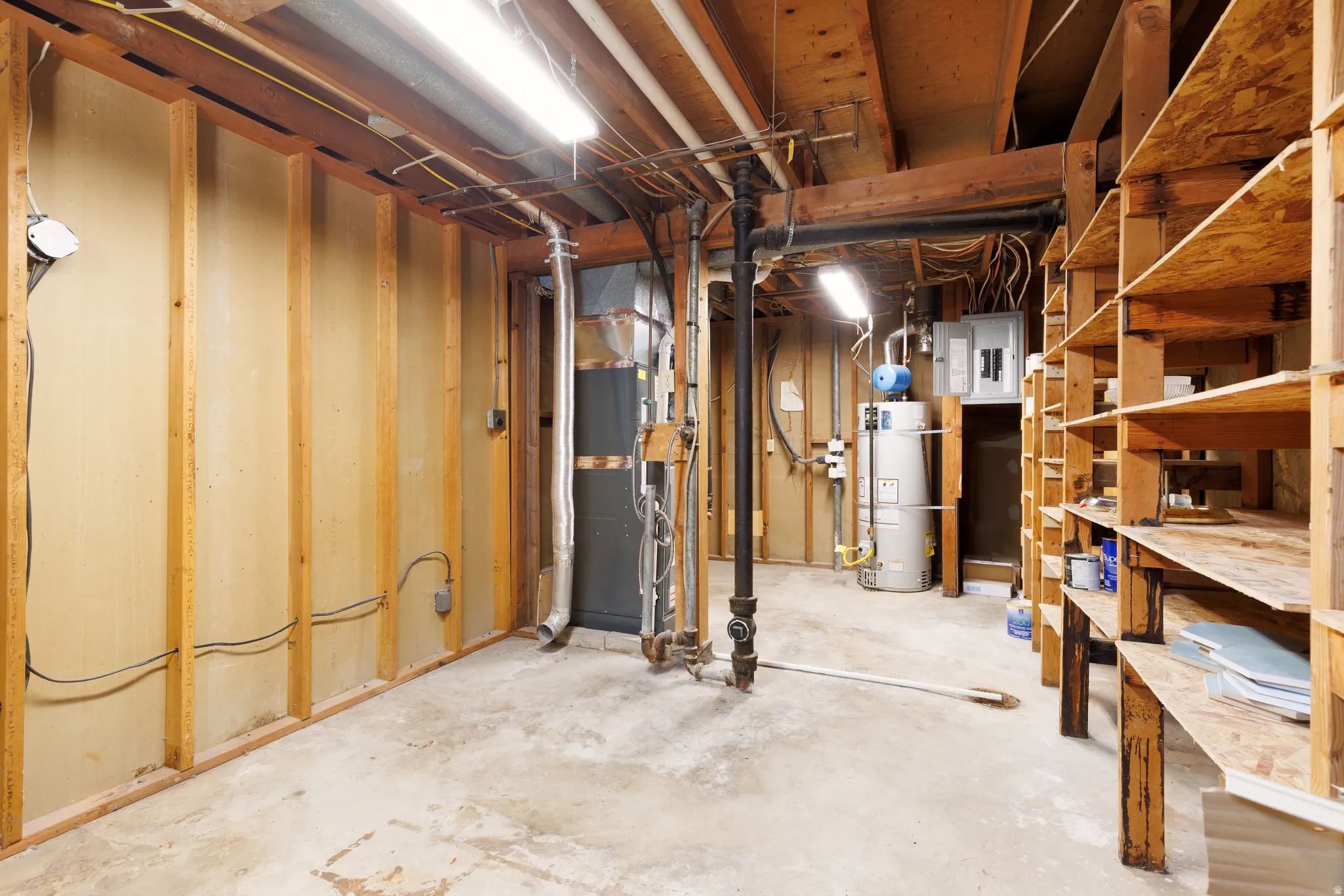 Spacious unfinished basement with exposed wooden framing, concrete flooring, fluorescent lighting, built-in wooden shelving, and visible utility systems including a water heater and electrical panel