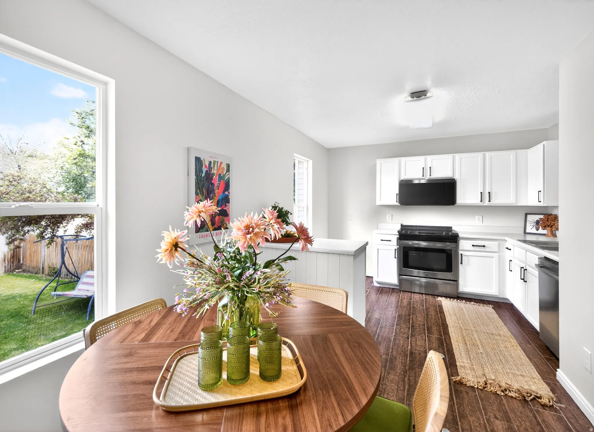 Bright kitchen and dining area featuring white cabinetry, stainless steel appliances, wood-finish flooring, and a large window overlooking the backyard