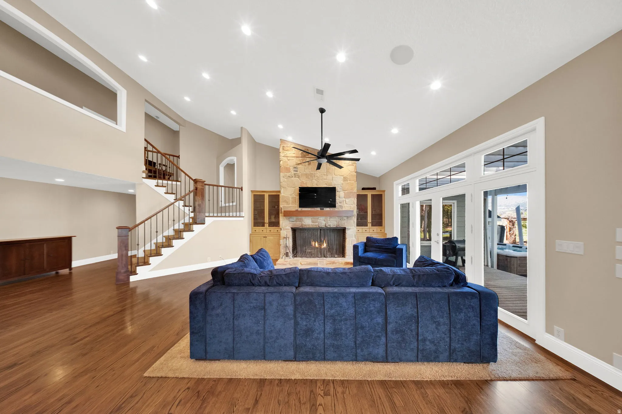 Spacious living area featuring high vaulted ceilings, recessed lighting, wood-finish flooring, a stone fireplace with a wood mantle, and built-in cabinetry, complemented by a wall of French doors with transom windows