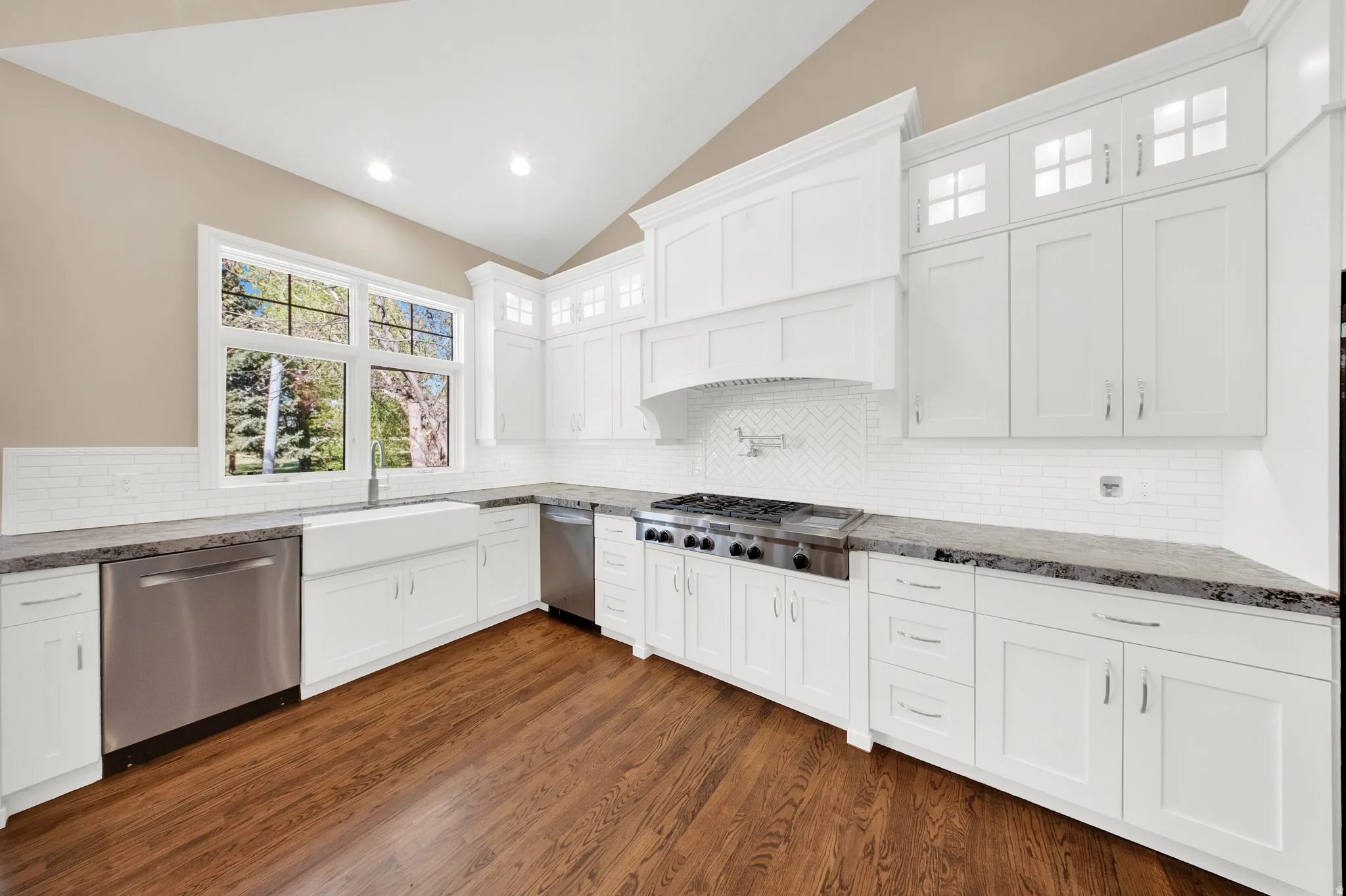 Kitchen featuring white shaker cabinetry with upper glass-front accents, stainless steel appliances, a farmhouse sink, and wood-finish flooring
