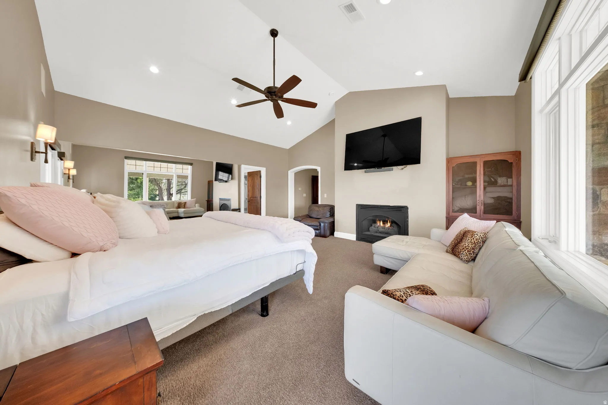 Spacious Primary bedroom featuring vaulted ceilings, a ceiling fan, sconce lighting, and a fireplace with a mounted television