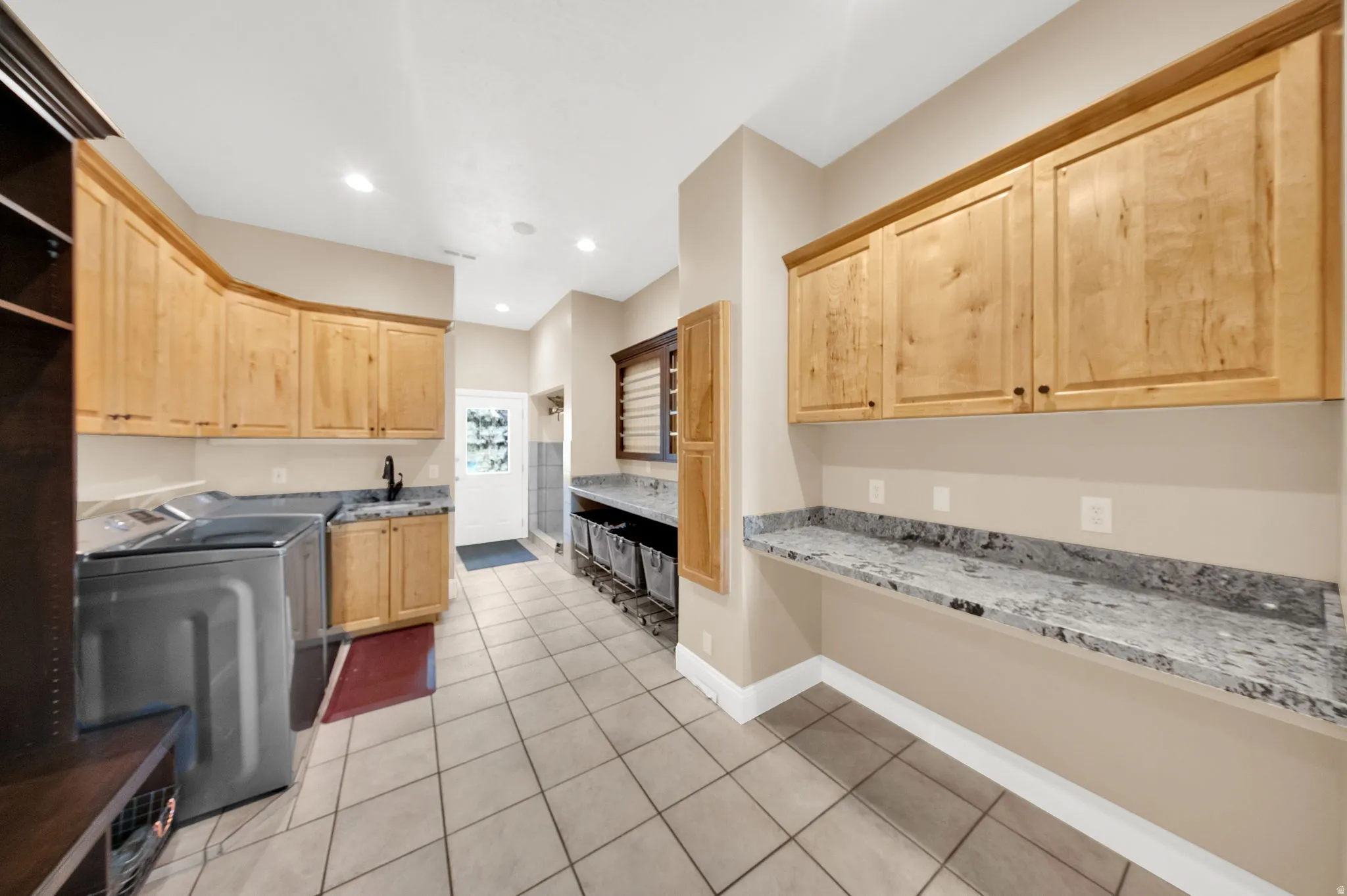 Spacious utility room featuring extensive light wood cabinetry, light-toned tile flooring, and ample counter space with a speckled stone-finish surface