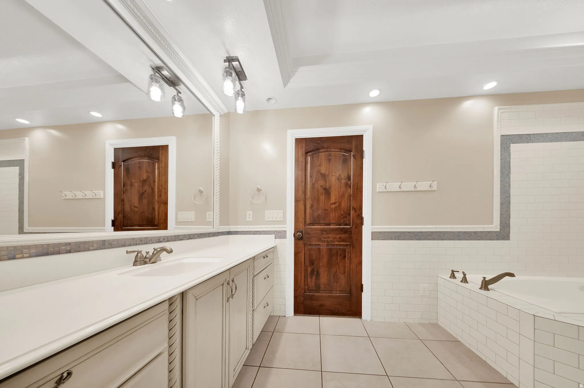 Bathroom featuring a large vanity with dual sinks, an expansive mirror, recessed lighting, a built-in tub with tile surround, and a dark wood-finish interior door