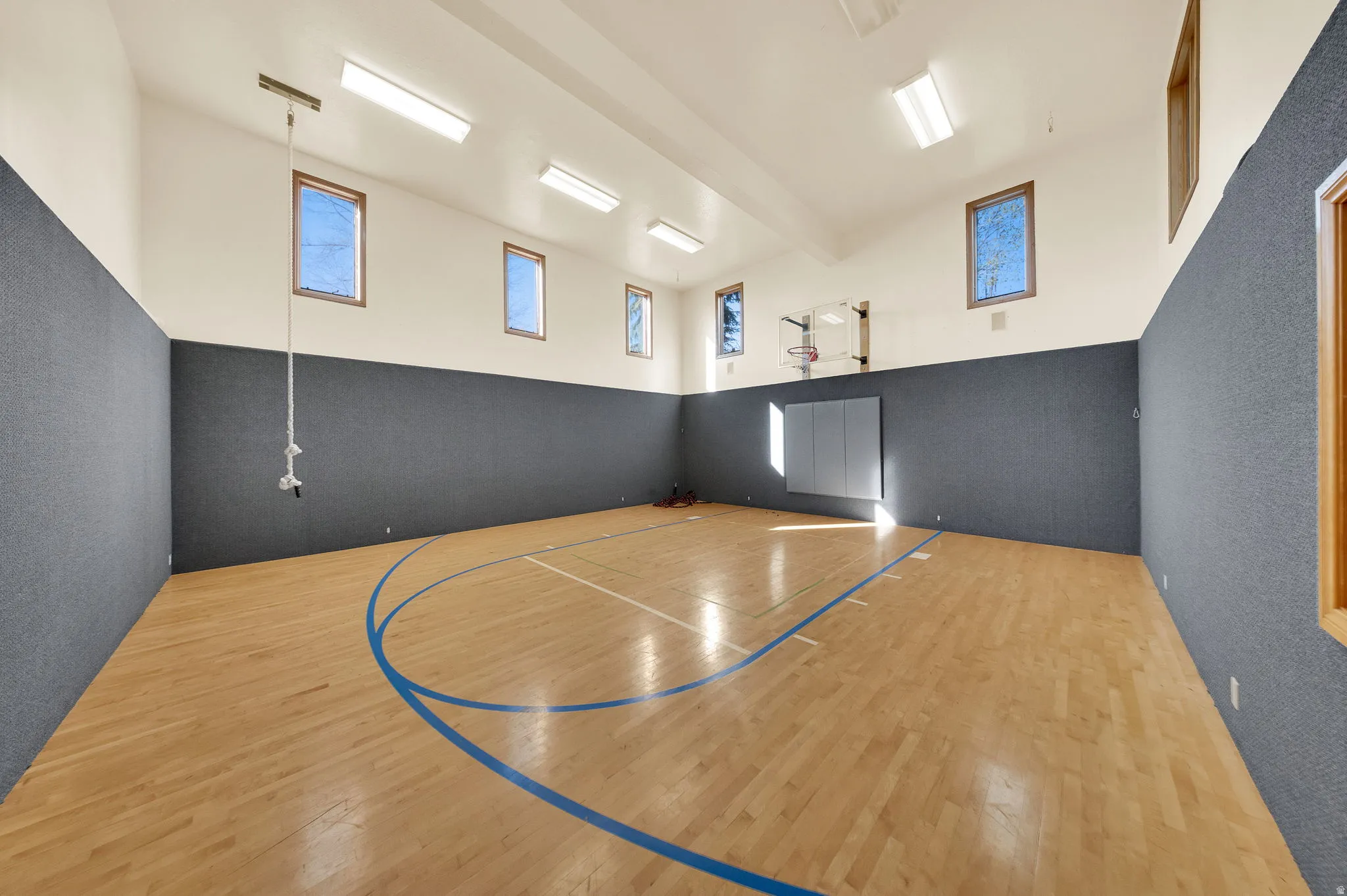 Indoor multi-sport court featuring wood-finish flooring with court markings, padded wall surfaces, a basketball hoop, and multiple windows