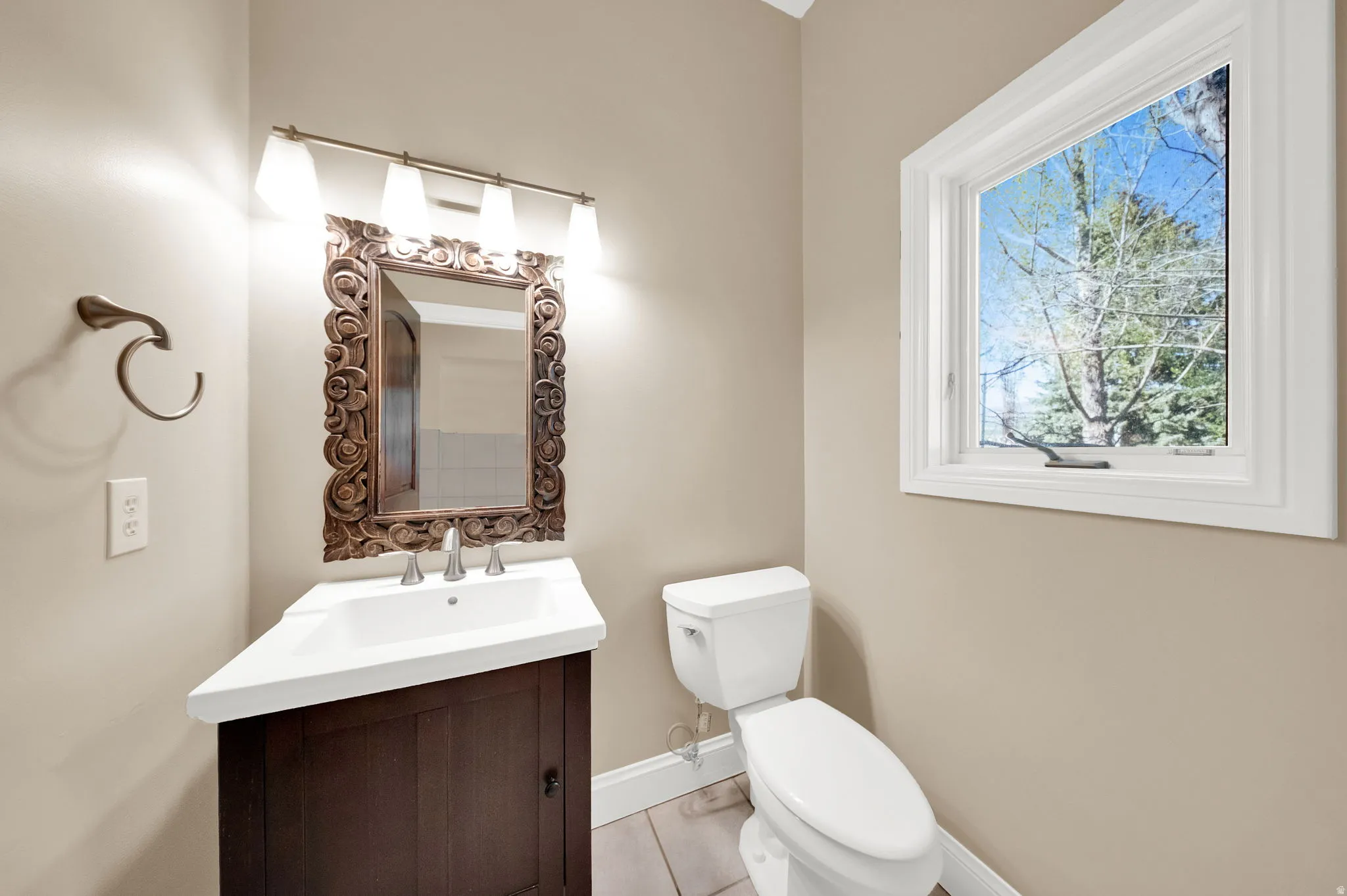 Bathroom featuring a dark wood vanity with white countertop, decorative carved wood-frame mirror, and a window with white trim