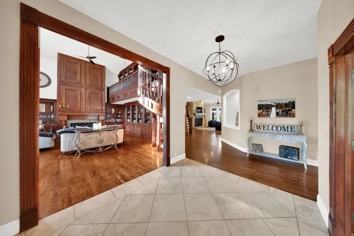 Foyer with tiled flooring and an ornate spherical chandelier, opening to a living area featuring wood-finish flooring, a built-in entertainment center with extensive cabinetry, a fireplace, and an elevated wood-paneled balcony with a carved railing