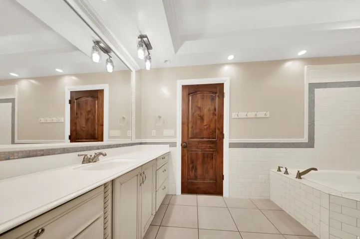 Bathroom featuring a large vanity with dual sinks, an expansive mirror, recessed lighting, a built-in tub with tile surround, and a dark wood-finish interior door