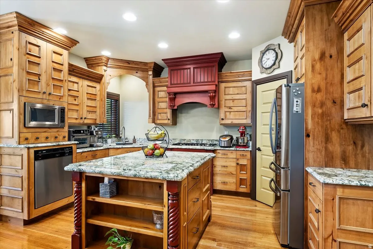 Kitchen featuring natural wood cabinetry, stone countertops, stainless steel appliances, a central island with carved accents, and wood-finish flooring