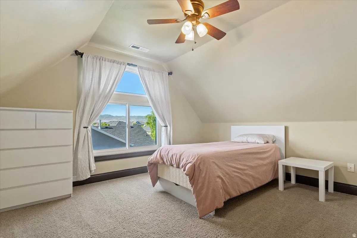 Room featuring a large window with white curtains, a ceiling fan with light fixture, neutral-toned carpeting, and sloped ceilings