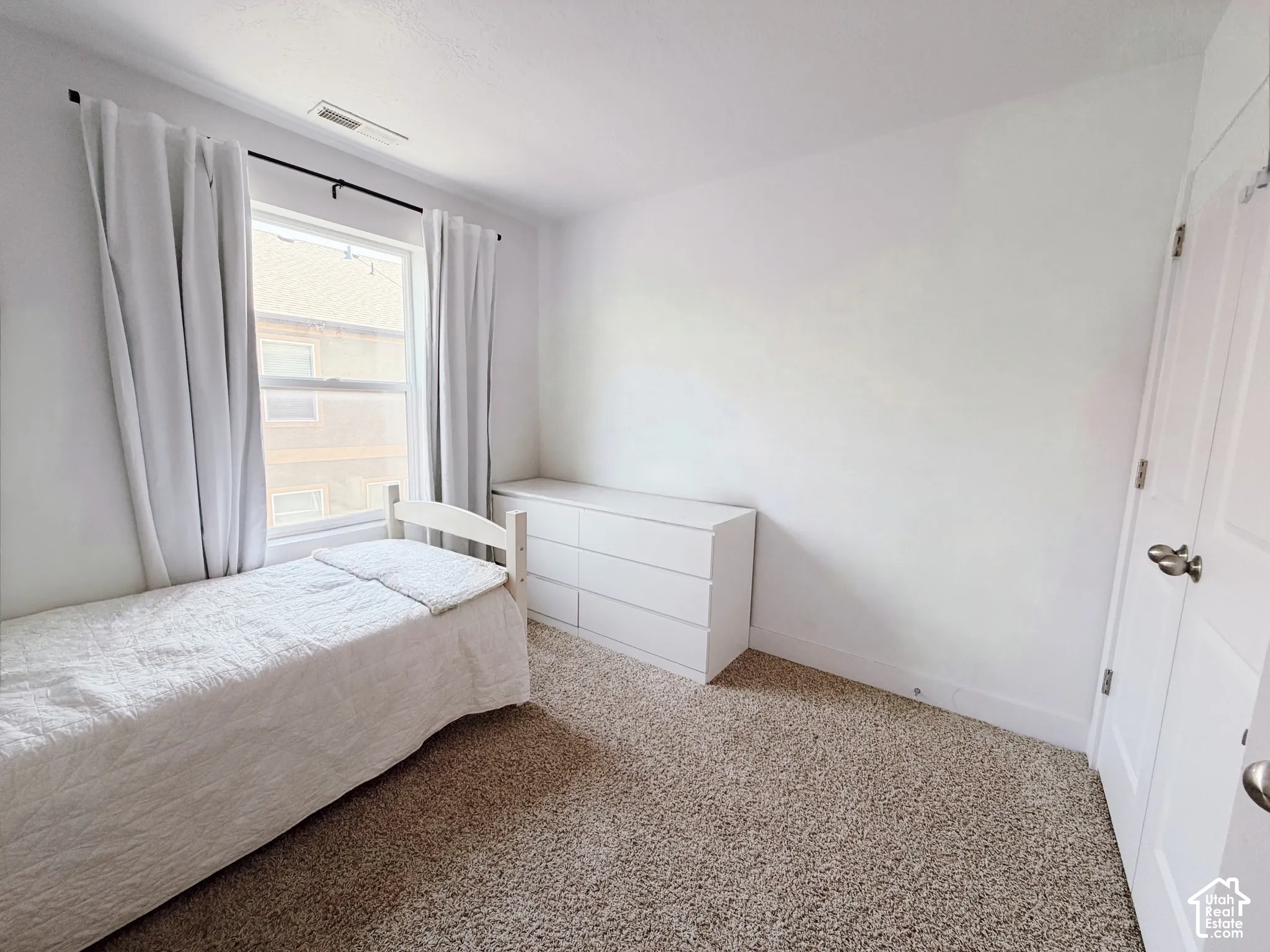 White walls, neutral carpet flooring, and a single window with curtain rod
