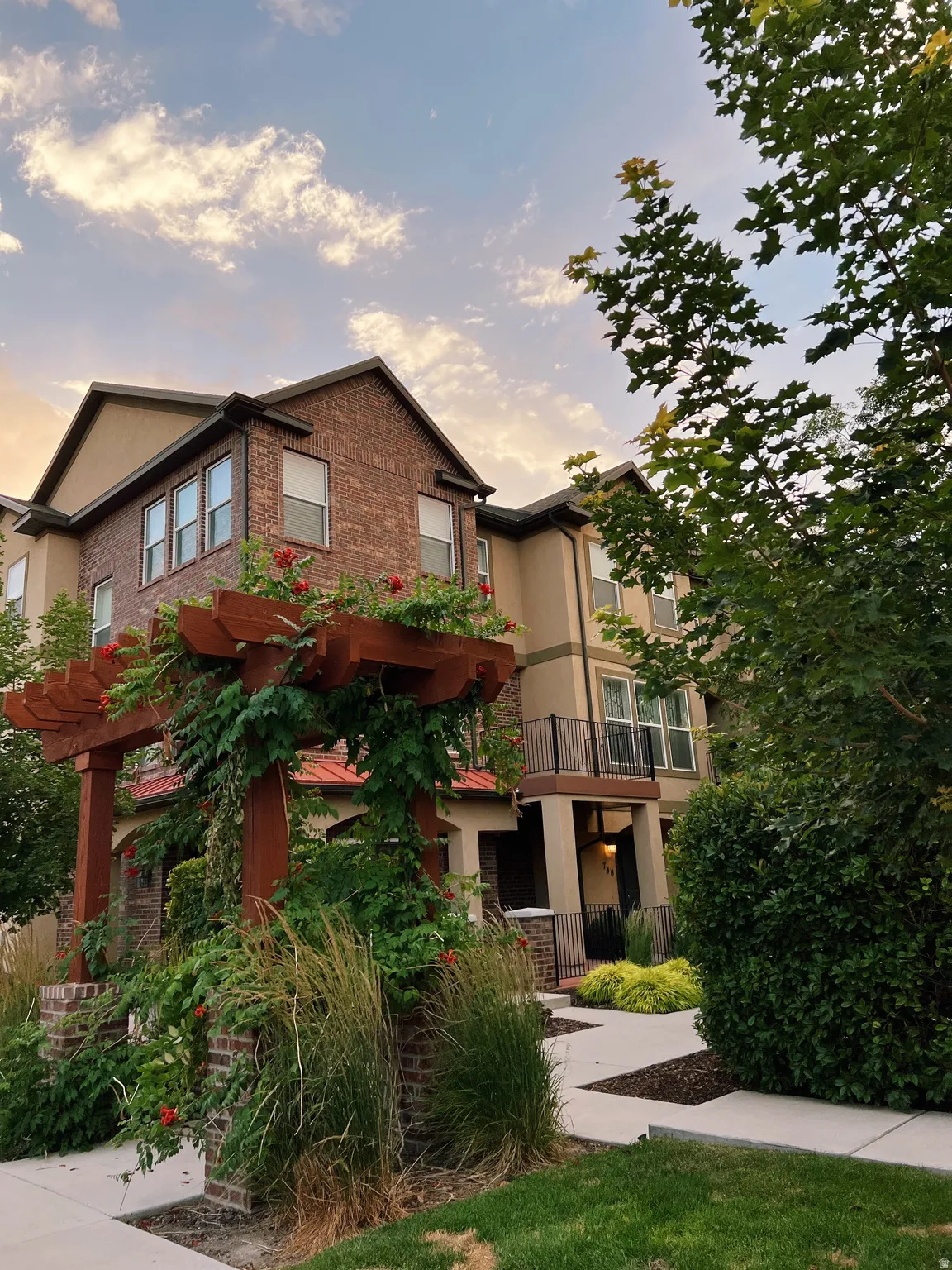 A decorative wooden pergola with climbing vines, a wrought iron balcony, a covered entryway, and mature landscaping