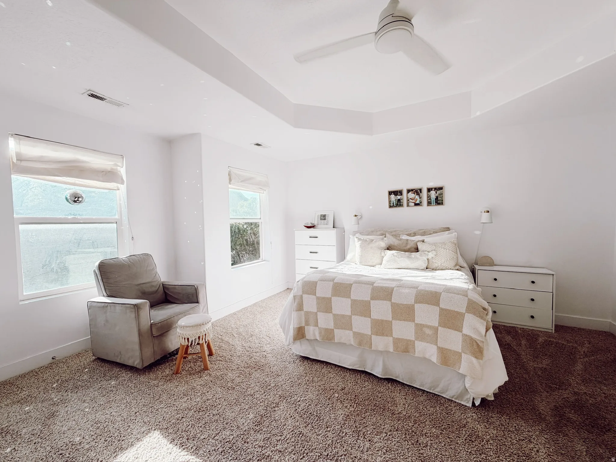 Spacious master bedroom featuring a tray ceiling, ceiling fan, two windows with window treatments, and wall-mounted sconces