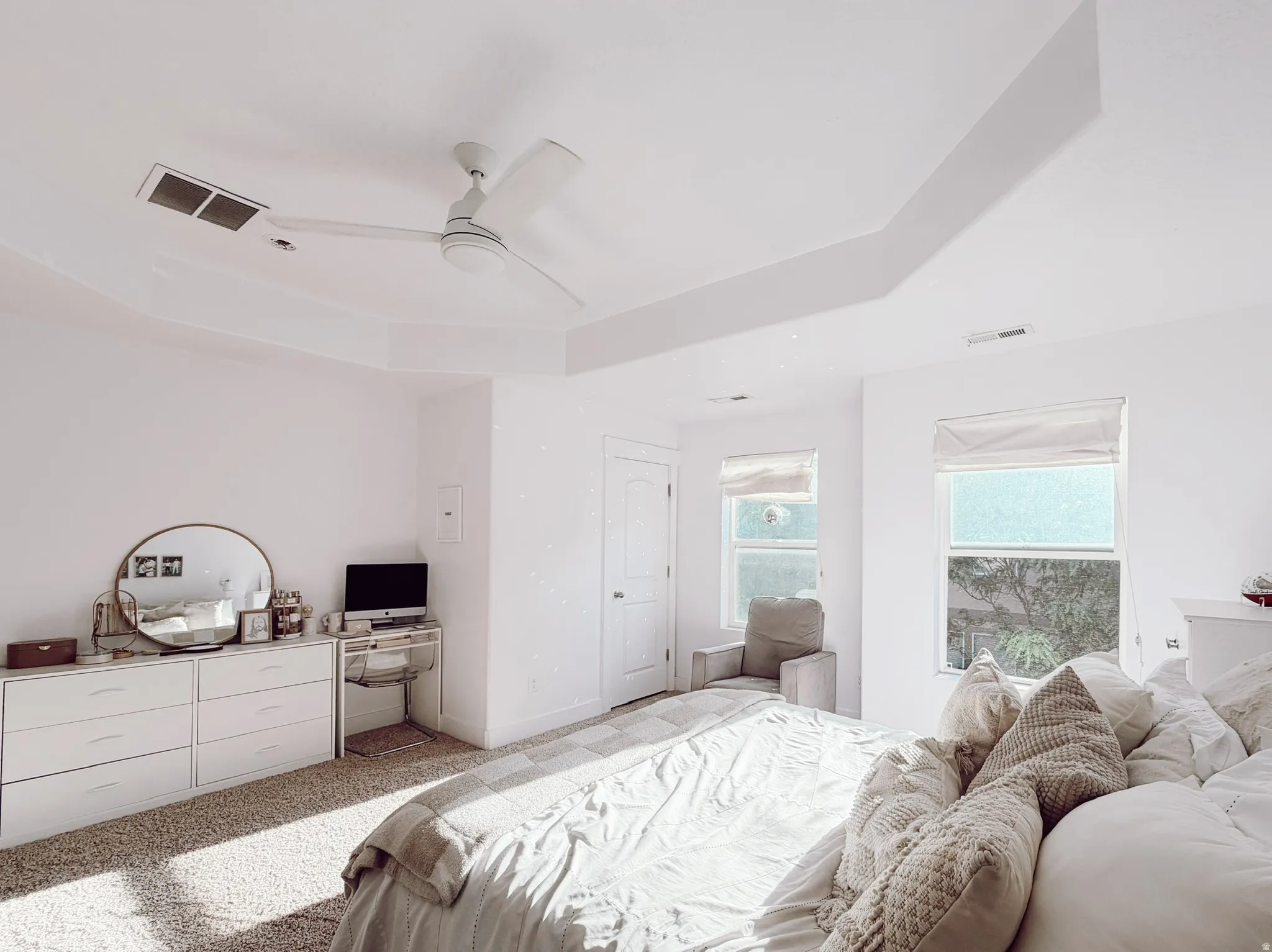 Spacious master bedroom featuring a tray ceiling with a modern ceiling fan, and two large windows with roller shades
