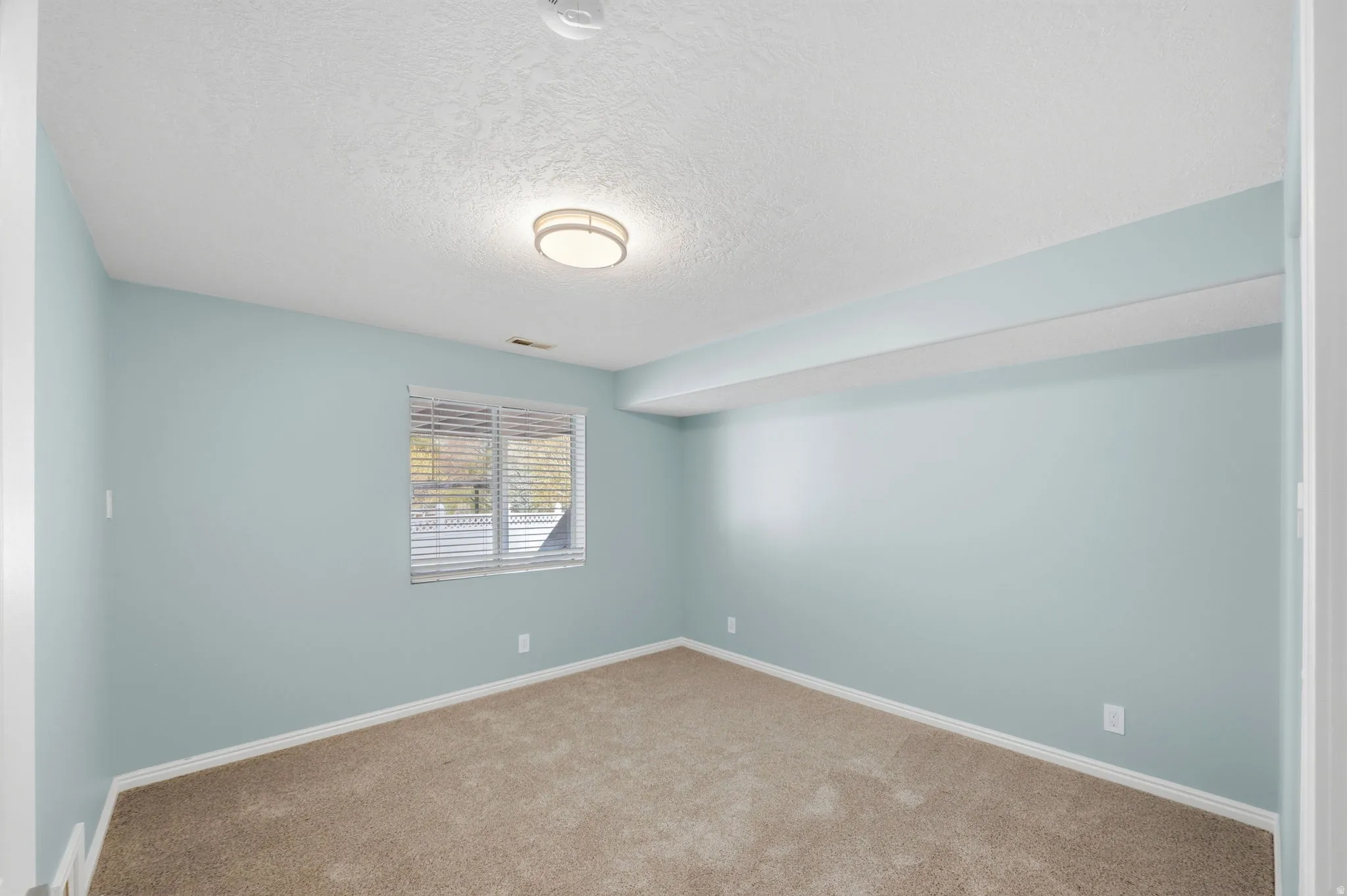 Spacious room featuring light blue walls, neutral carpeting, white baseboards, and a window with blinds